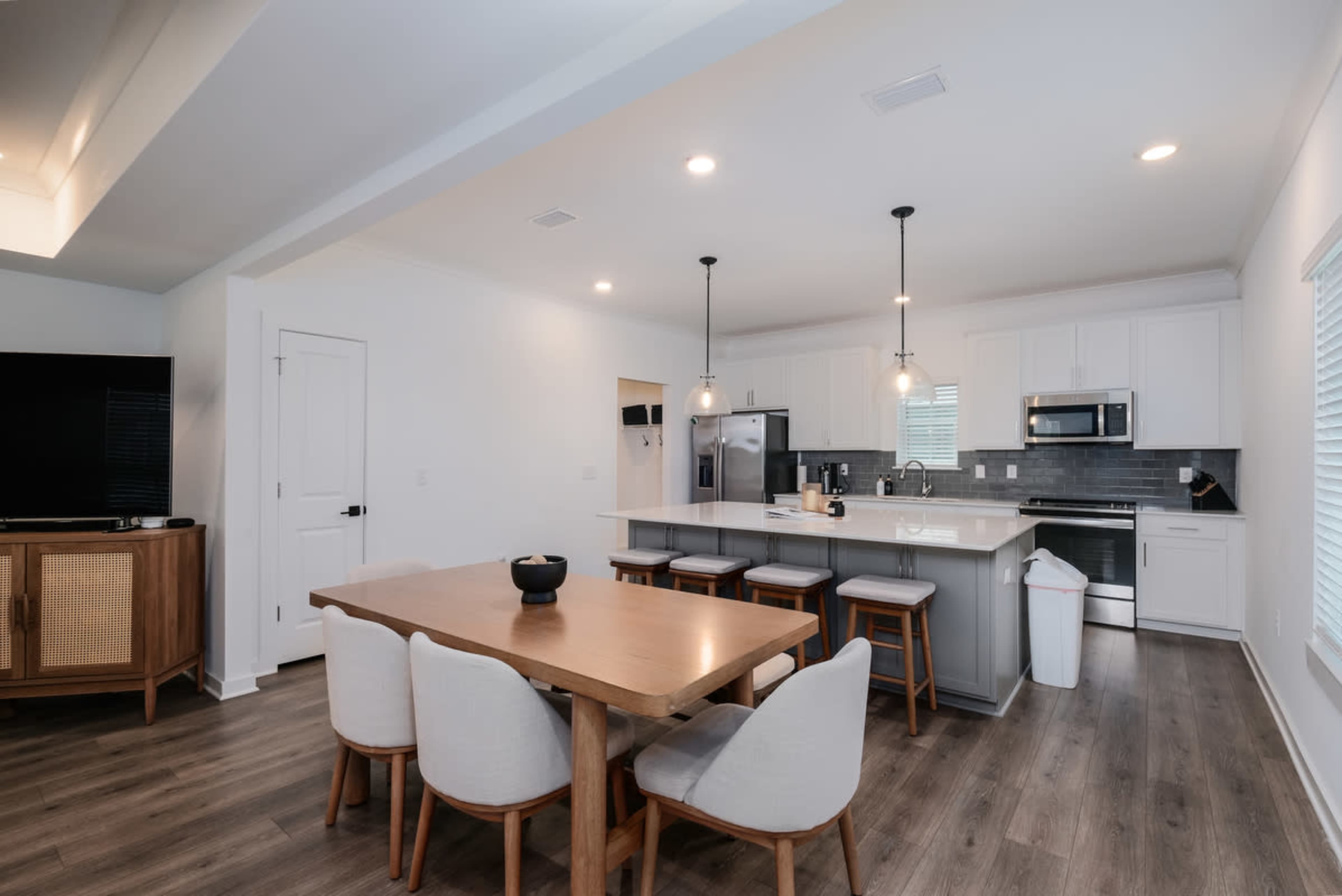 The image shows a modern kitchen and dining area featuring a wooden dining table with six chairs, a kitchen island with barstools, stainless steel appliances, and white cabinetry.