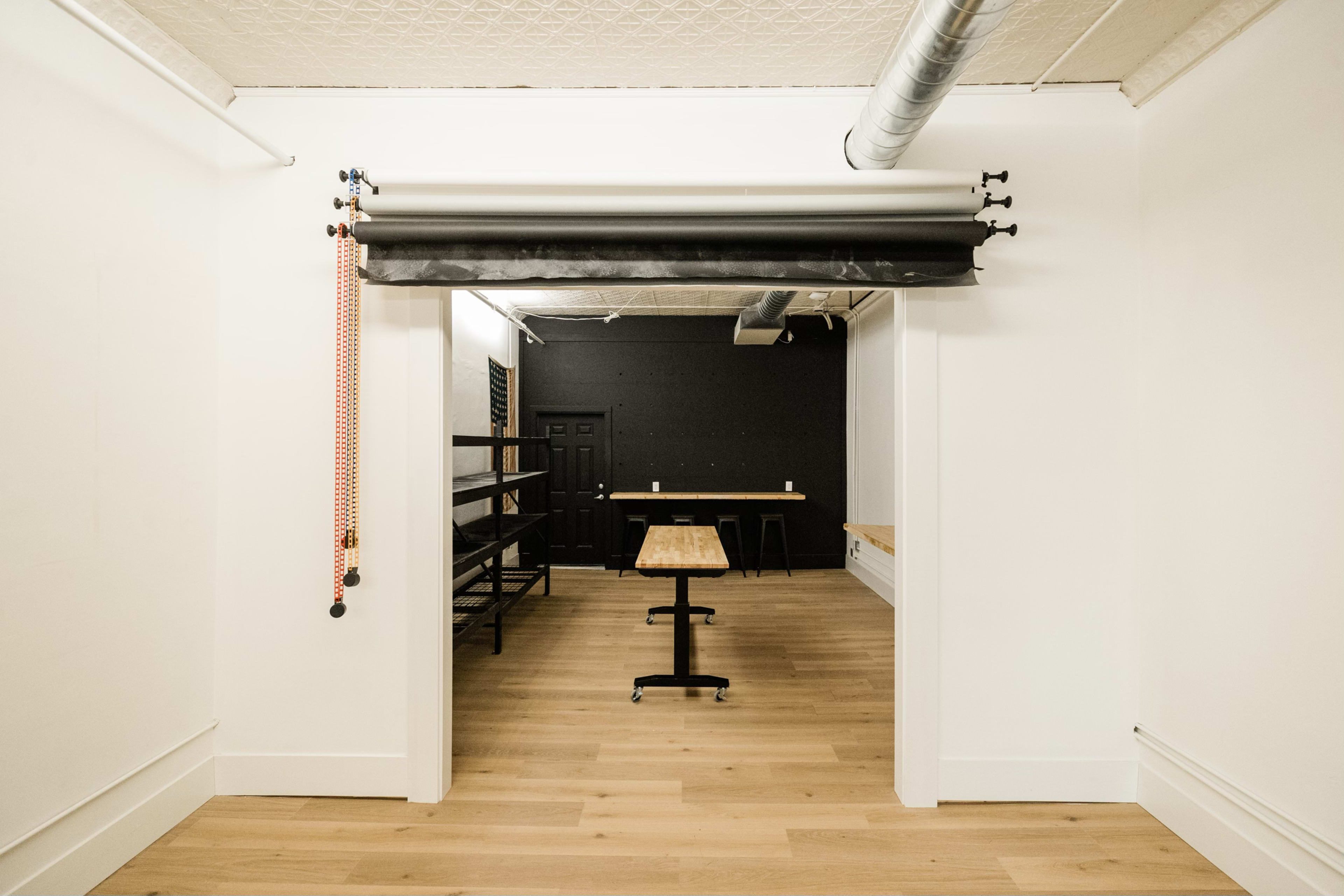 The image shows a minimalist interior space with a wooden table in the center, framed by a roll-up curtain at the entrance.