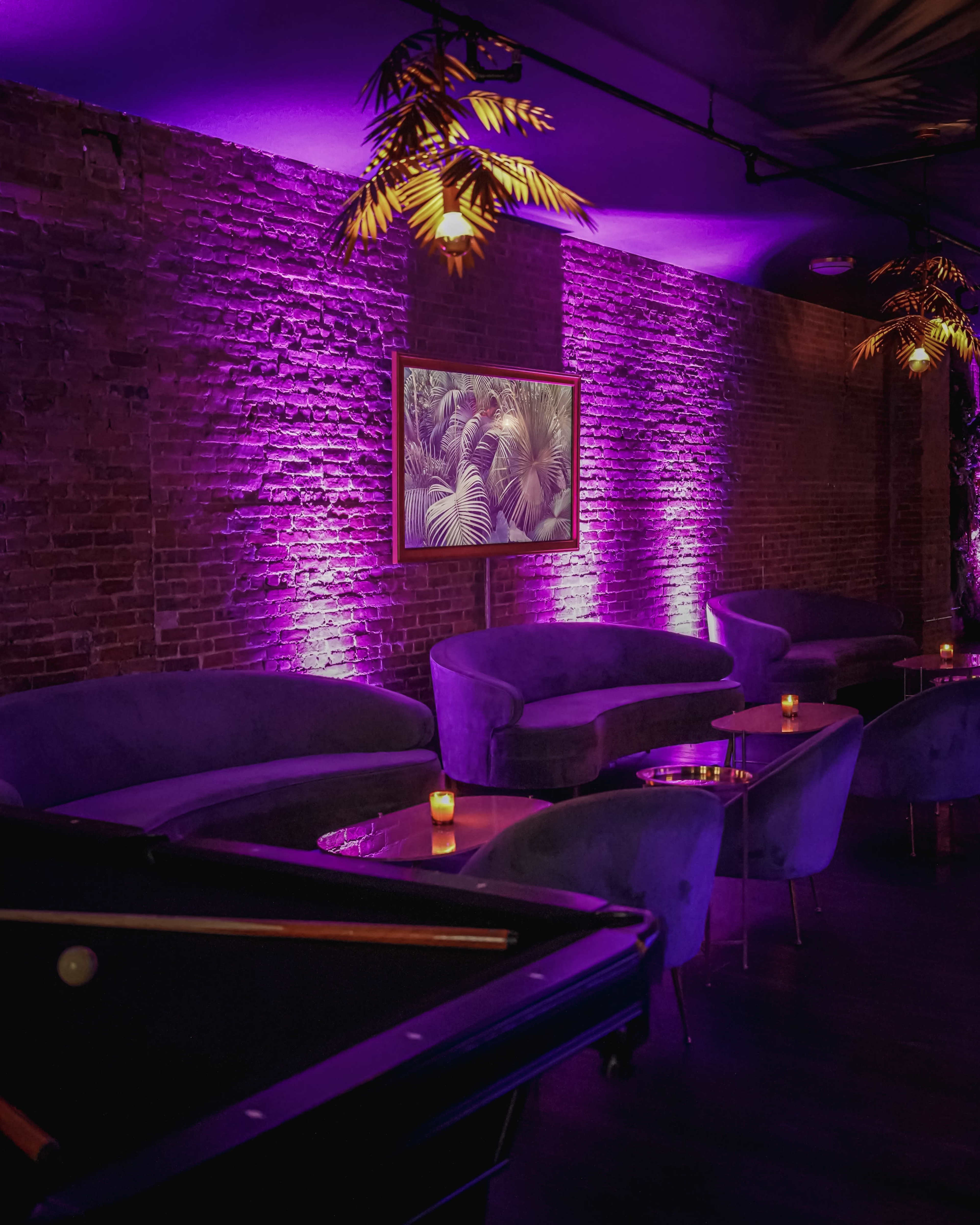 The image shows a dimly lit lounge space with purple lighting, featuring plush seating, a framed artwork of palm leaves, and small candles on tables.
