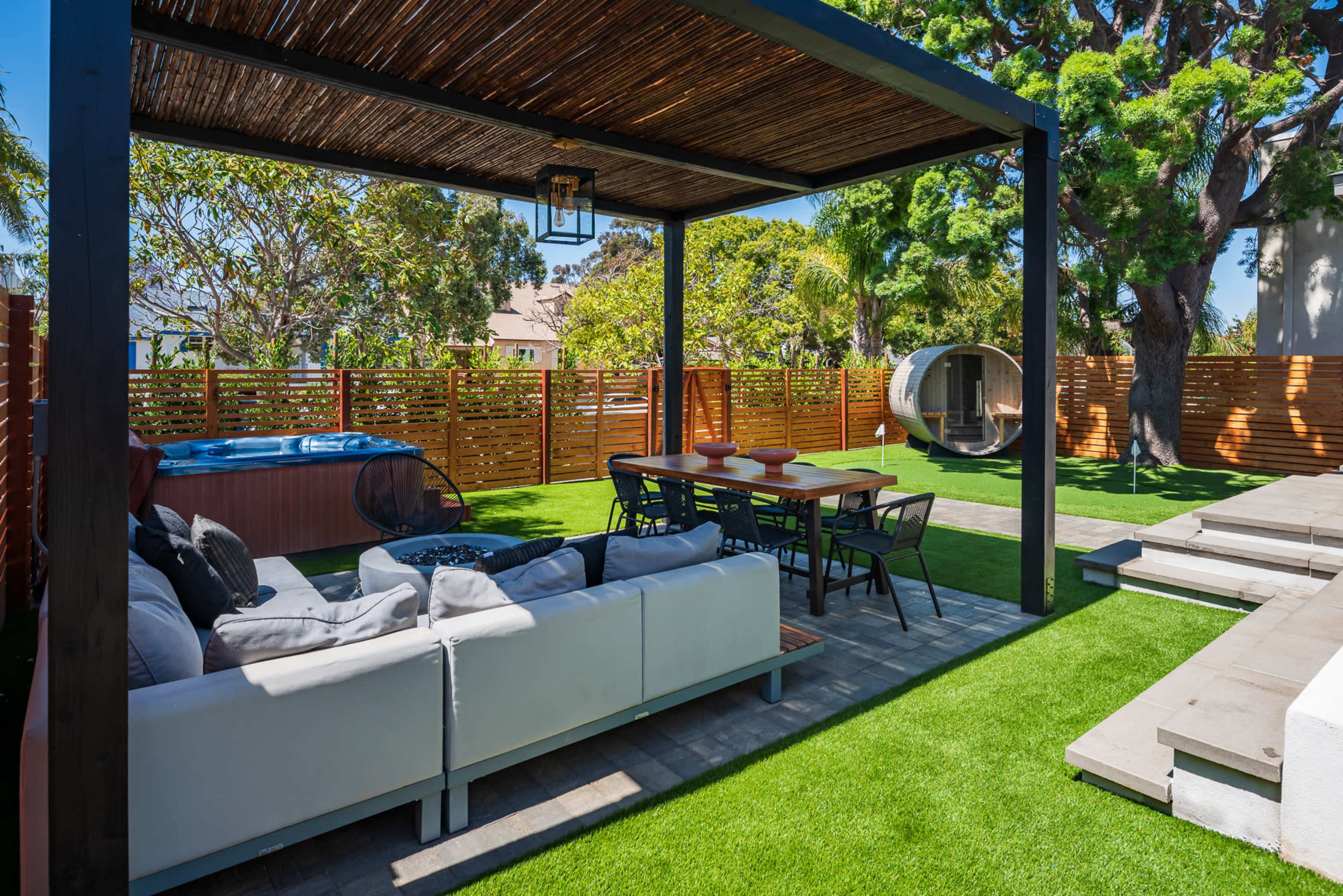 The image shows a modern outdoor patio with a seating area, dining table, and a small round structure, all surrounded by greenery and wooden fencing.