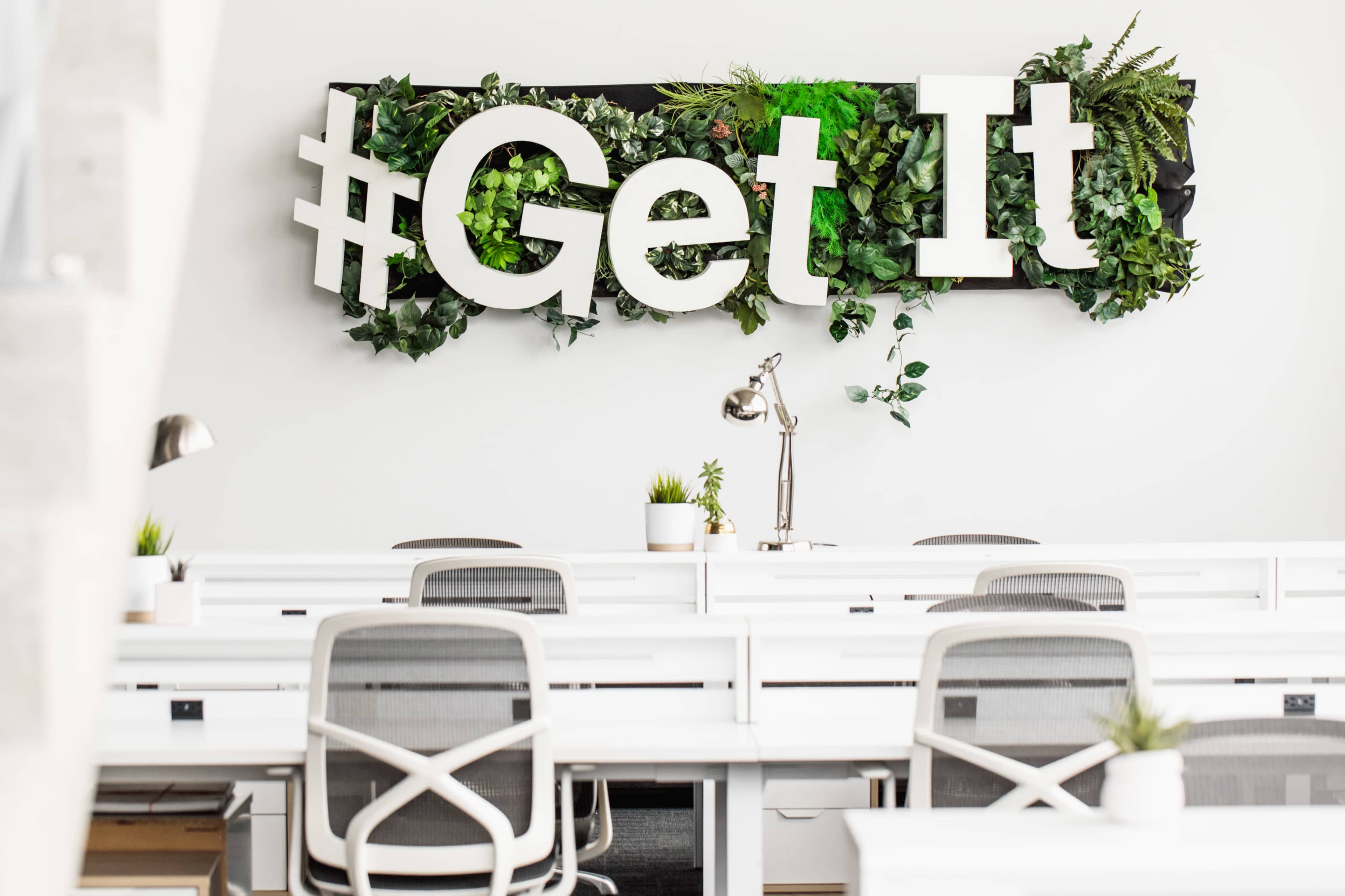 A modern office space featuring white desks and a decorative wall with the hashtag "#Get It" made of large letters surrounded by greenery.