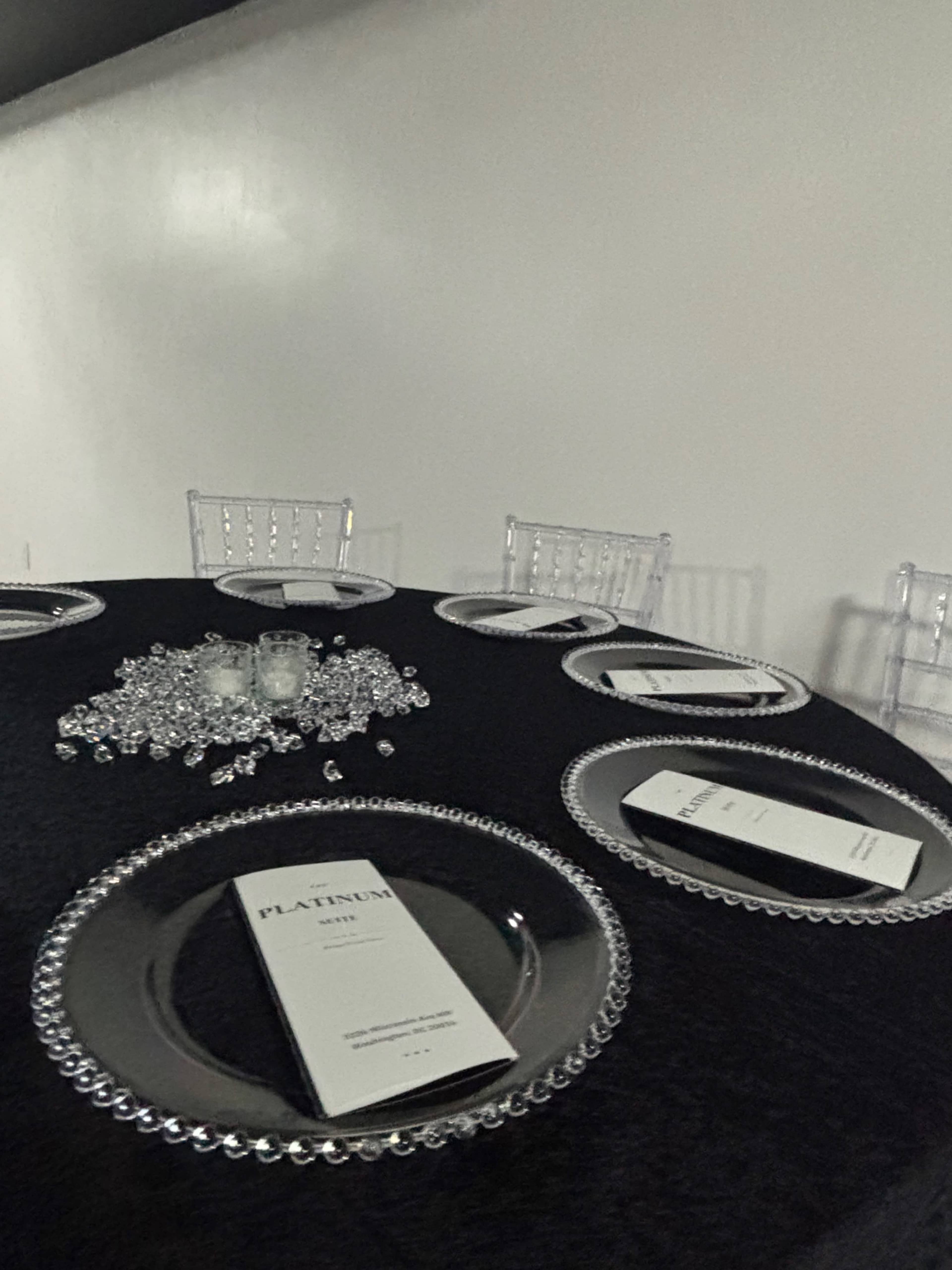 A round table is set with elegant black cloth, clear chiavari chairs, and decorative place settings featuring silver chargers and menus.