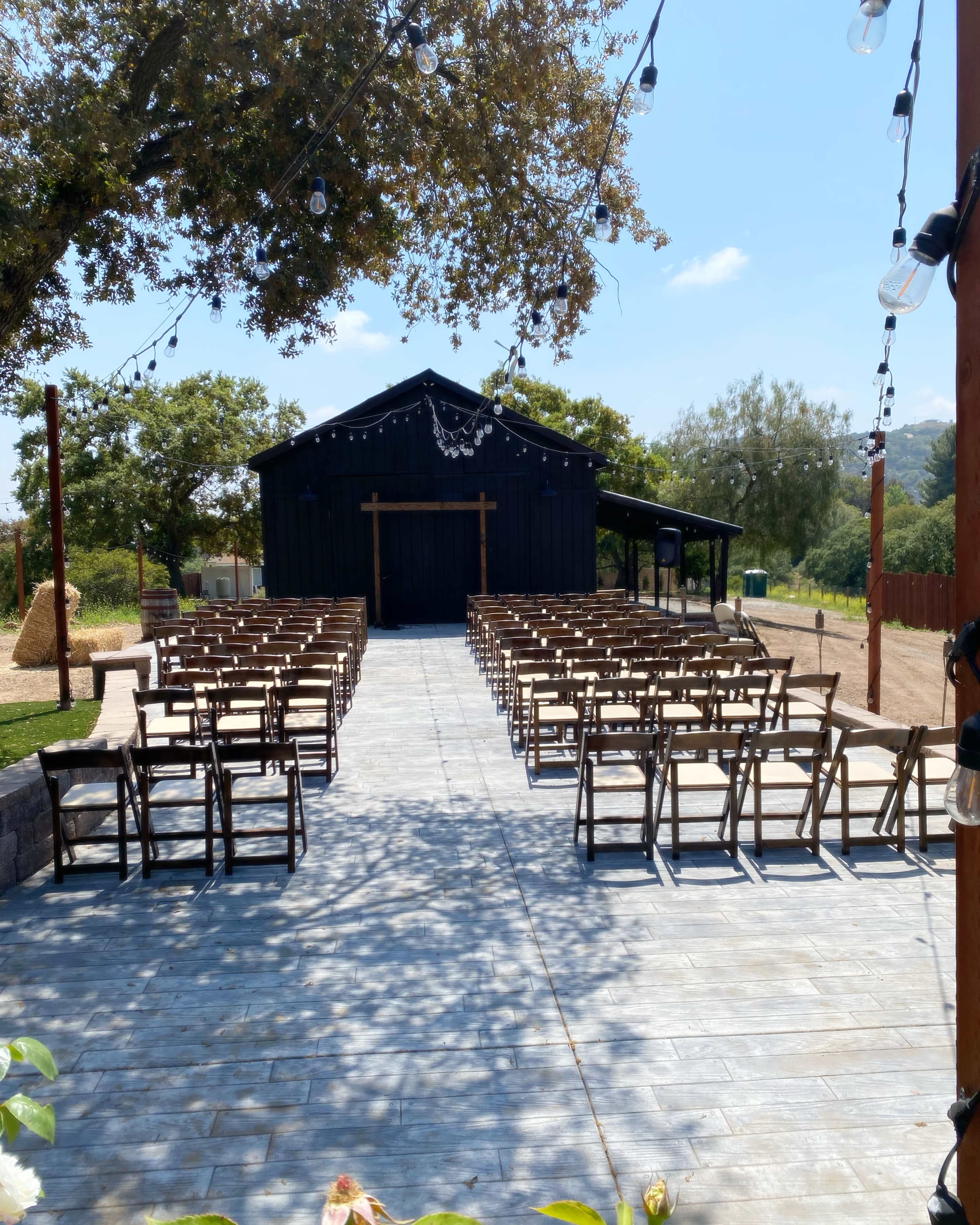 A pathway lined with wooden chairs leads to a dark wooden barn in a casual outdoor setting.