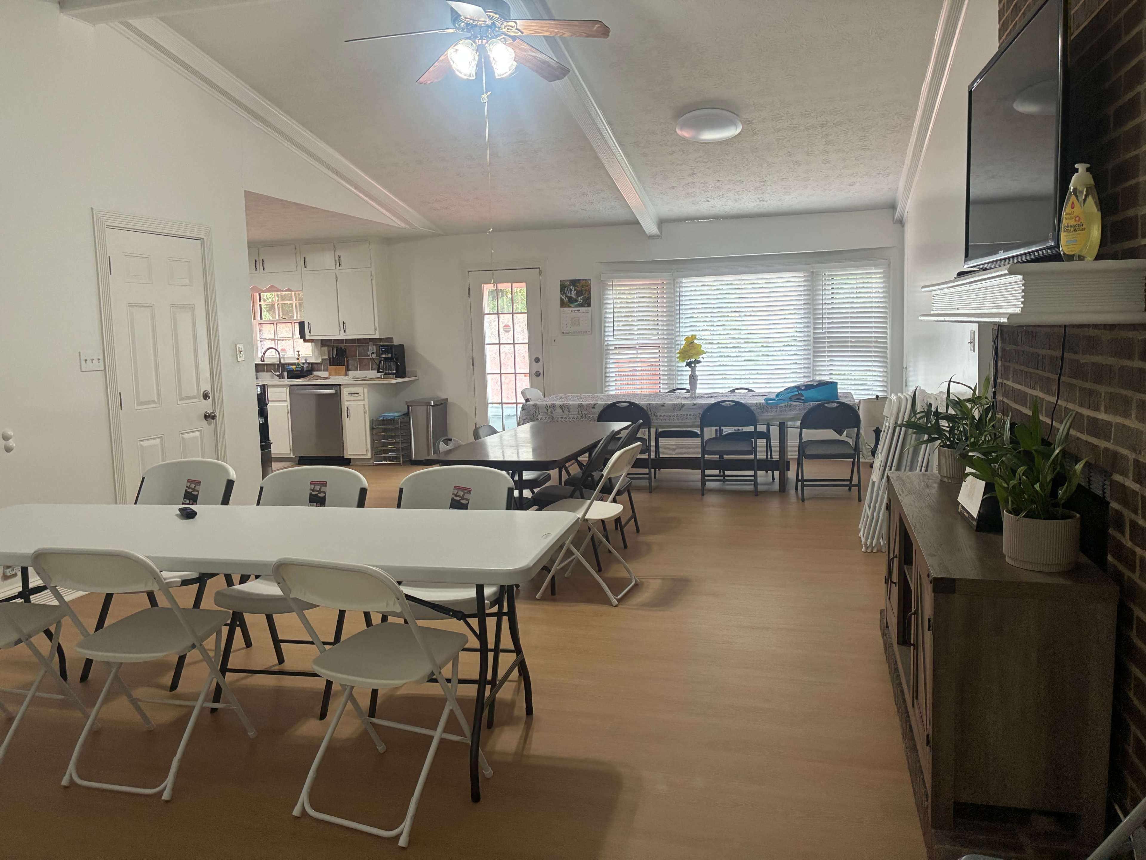 The image shows a spacious room with white walls, wooden flooring, several folding tables and chairs arranged, and a kitchen area visible in the background.