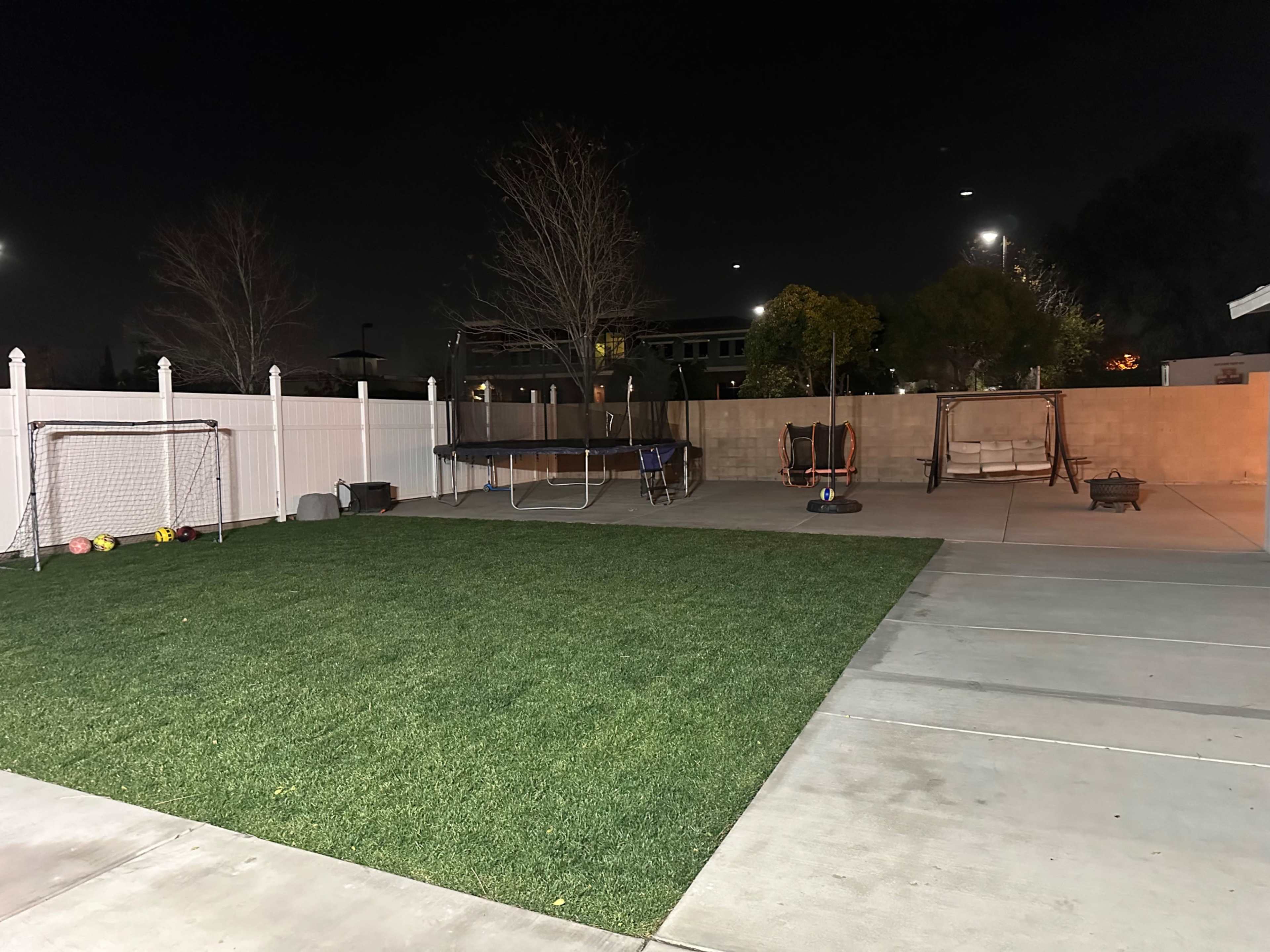 The image depicts a backyard at night featuring a grass area, a trampoline, two soccer goals, a swing set, and concrete patio sections.