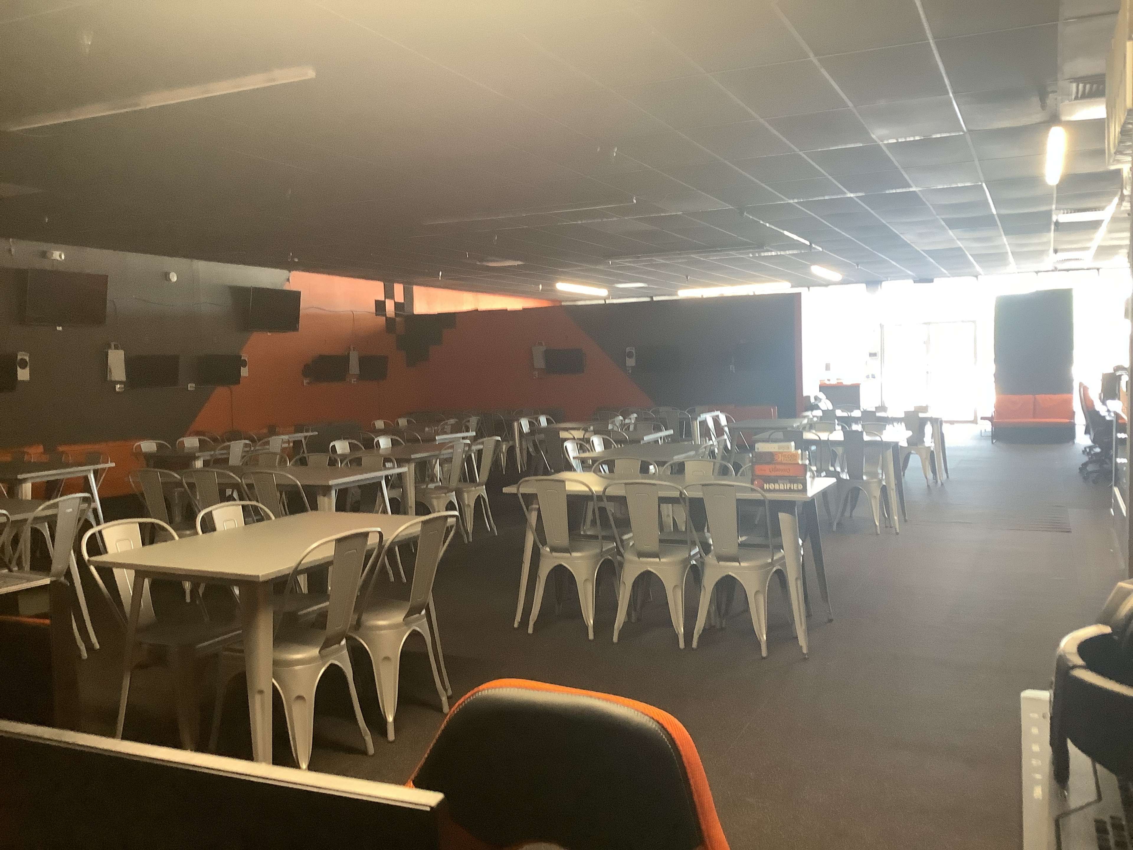 The image shows an empty dining area with metal tables and chairs, lined up in rows under dim lighting.