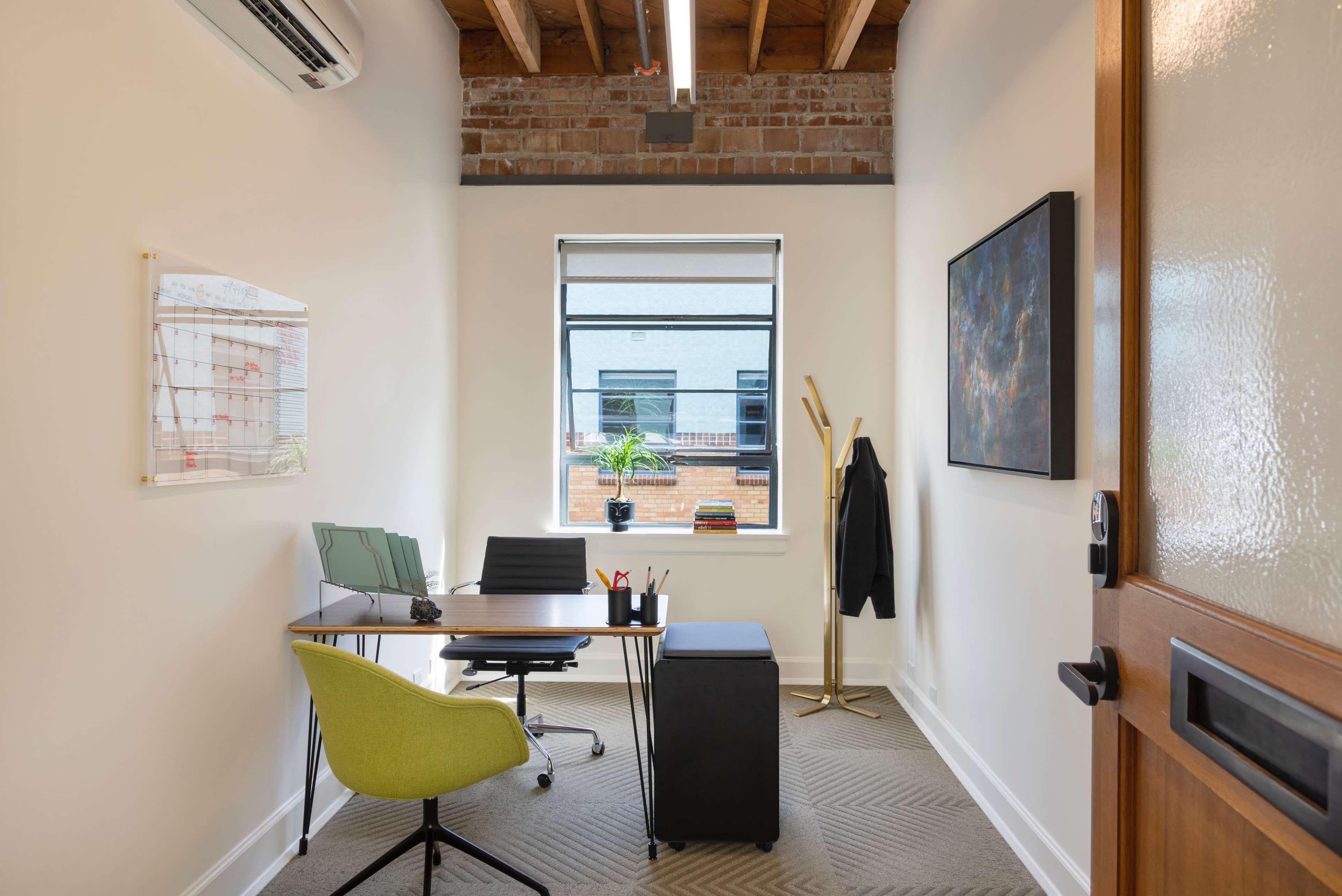 The image shows a small office featuring a desk with a green chair, a window with a view of a brick building, and a coat rack in the corner.