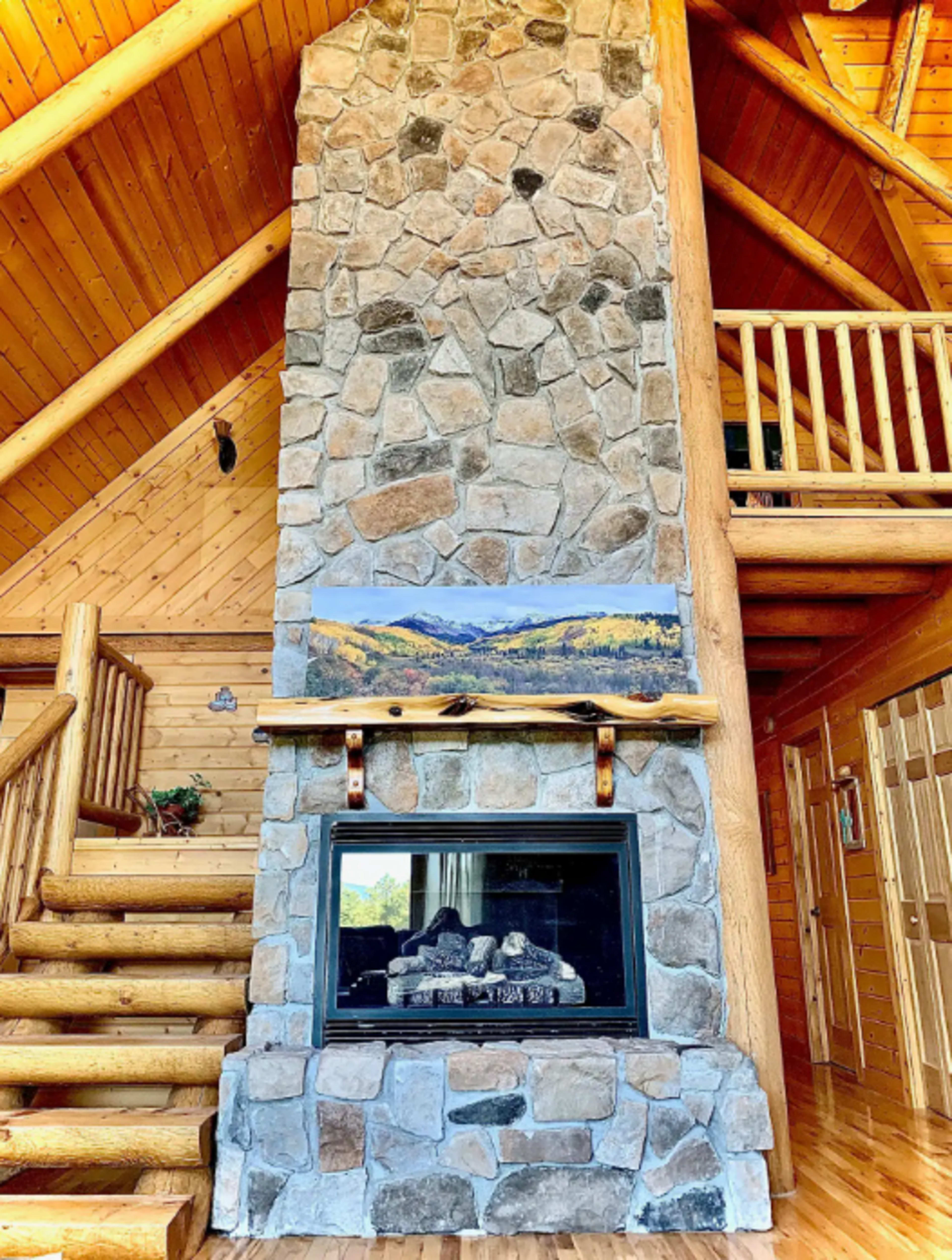 The image shows a large stone fireplace in a wooden cabin, with a staircase beside it and a landscape painting above the mantel.