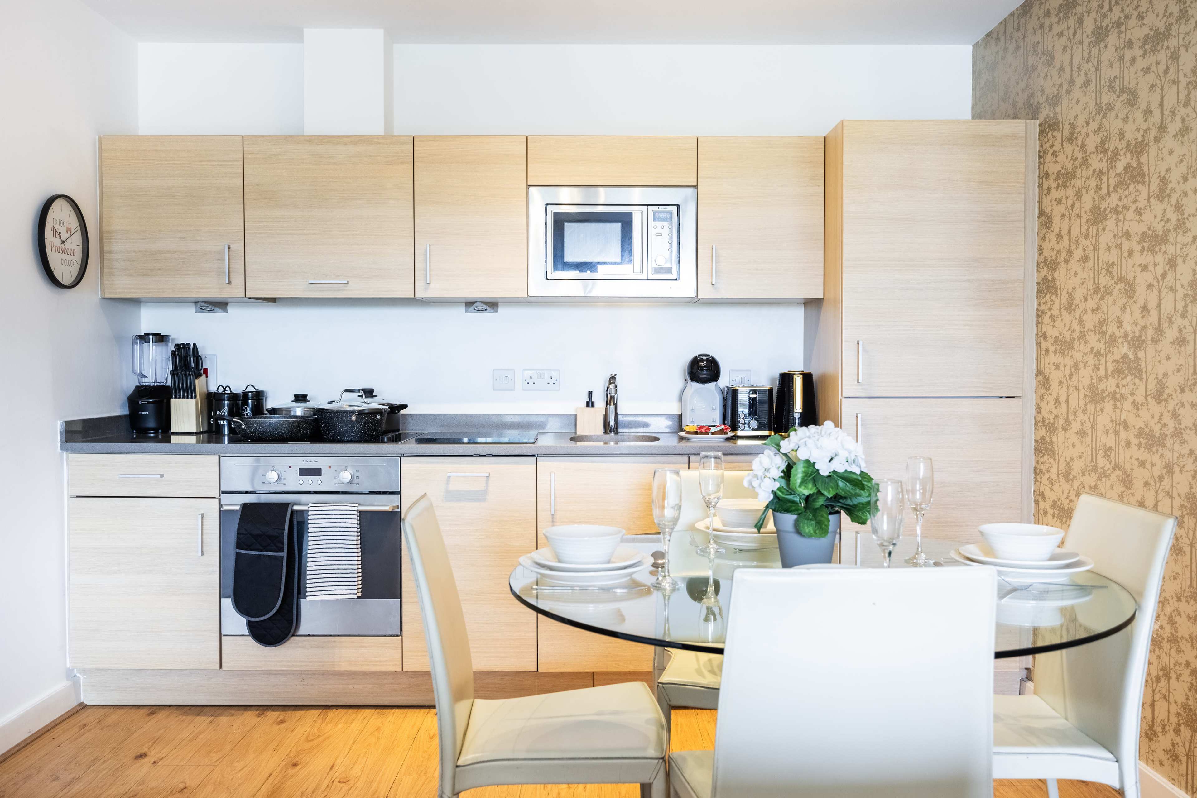 A modern kitchen featuring wooden cabinets, a built-in microwave, and a round glass dining table set for four.