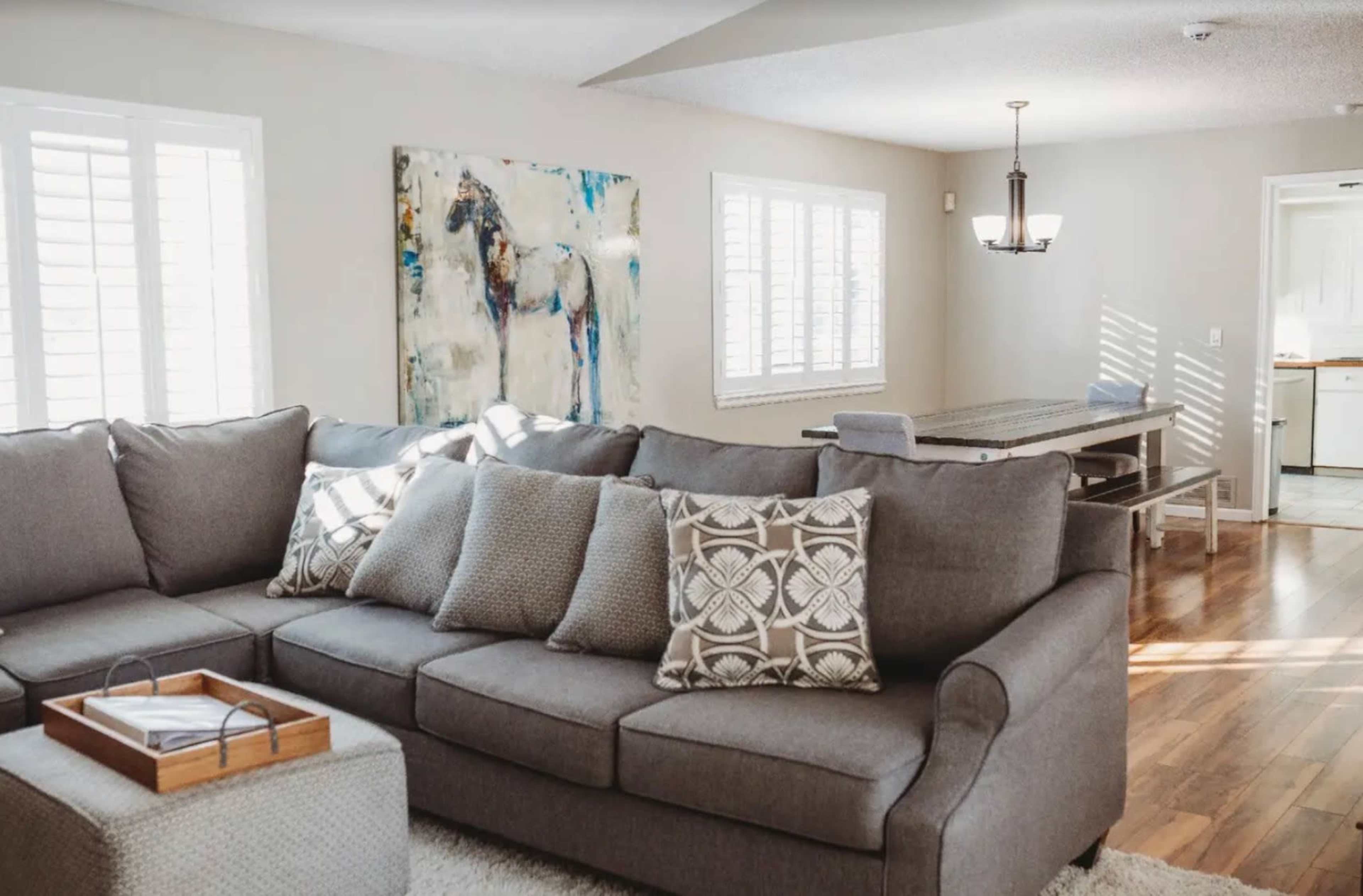 A living room features a gray sectional sofa with patterned pillows, an artwork on the wall, and a dining table in the background.