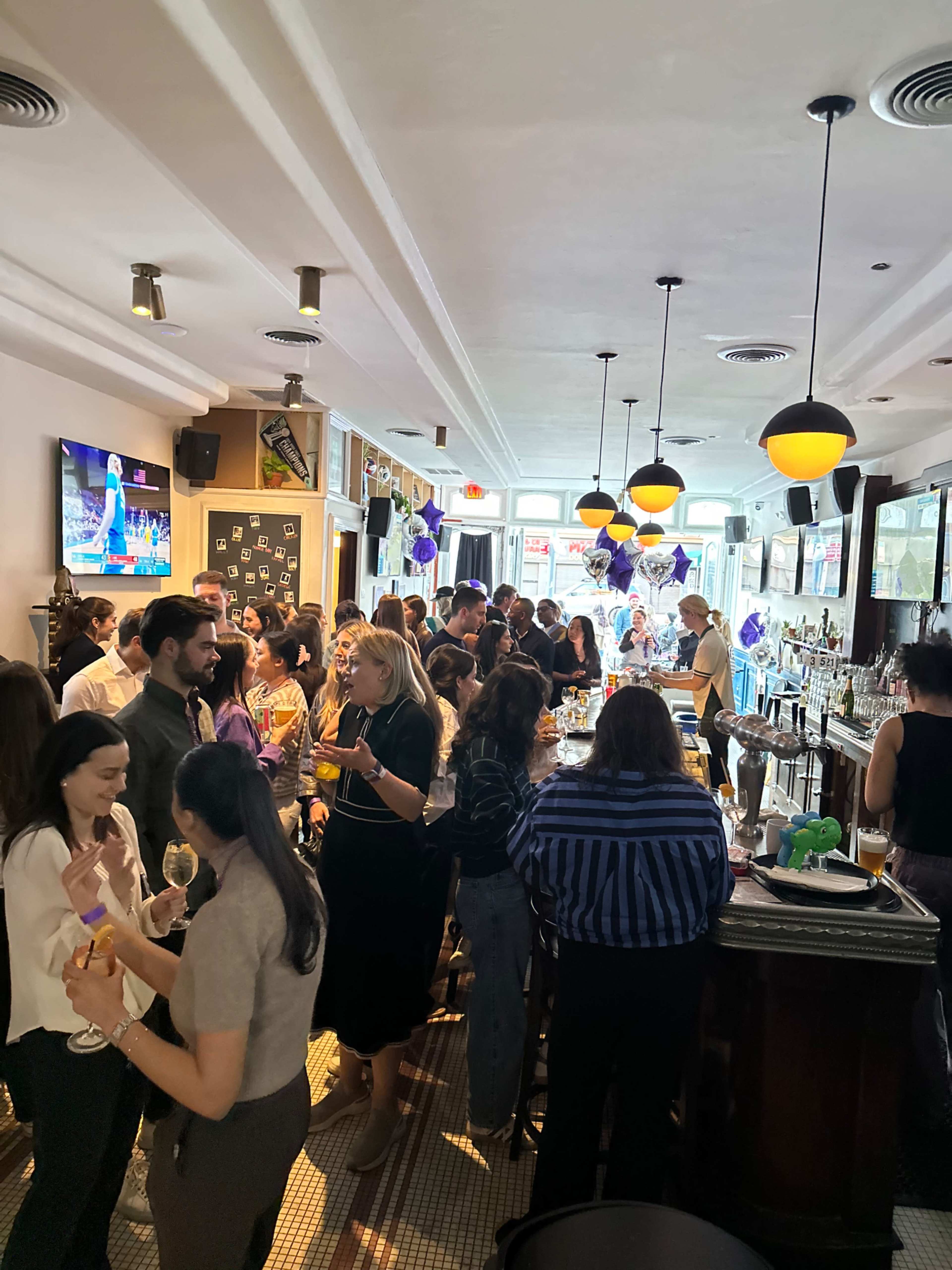 A crowded bar scene features a diverse group of people socializing, with drinks in hand and a television displaying a sports event in the background.