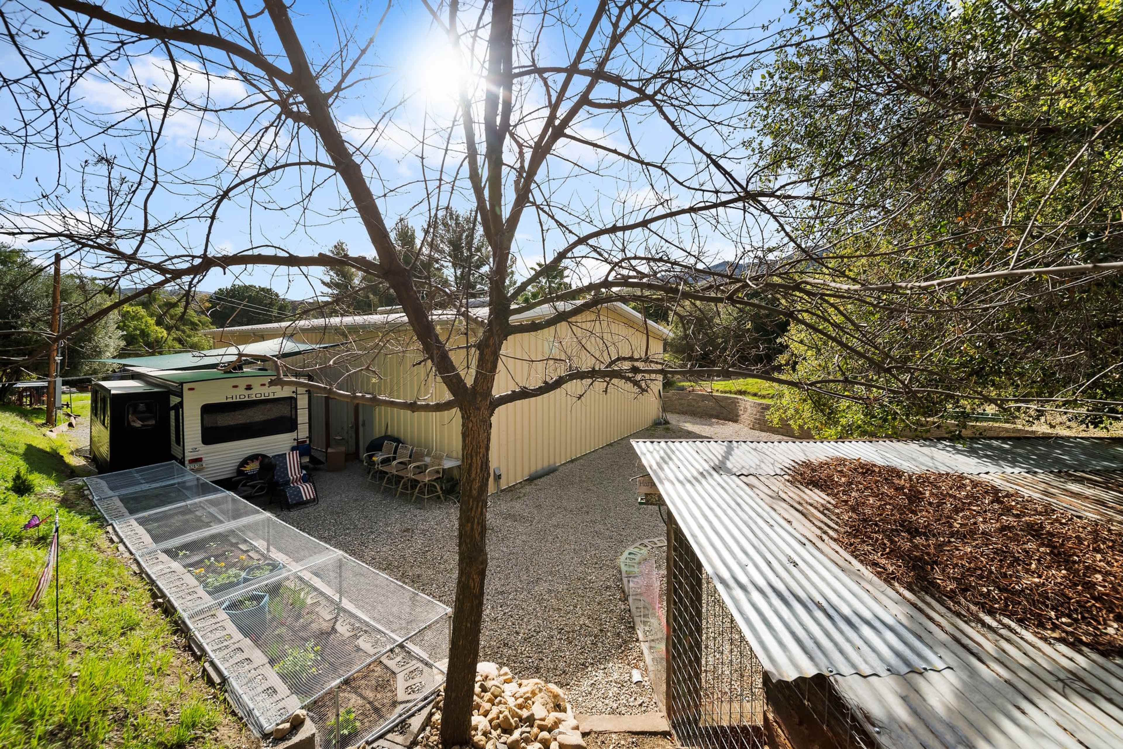 The image shows a rural scene featuring a yellow building surrounded by gravel, a nearby trailer, and a partially covered area with a tree in the foreground.