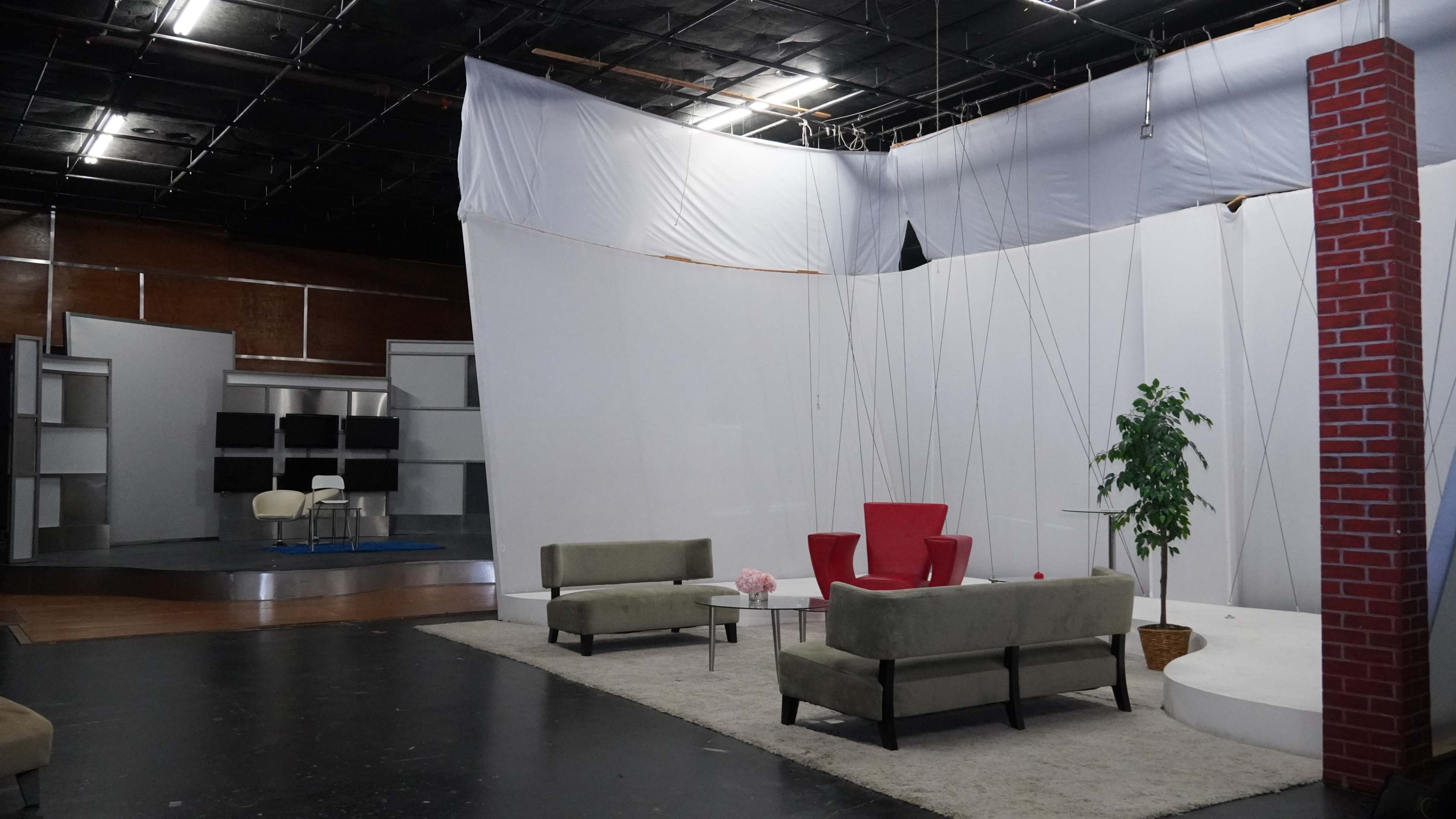 The image shows a television studio set featuring a white backdrop, two green sofas, a round table with a red chair, and a potted plant on a carpeted area.