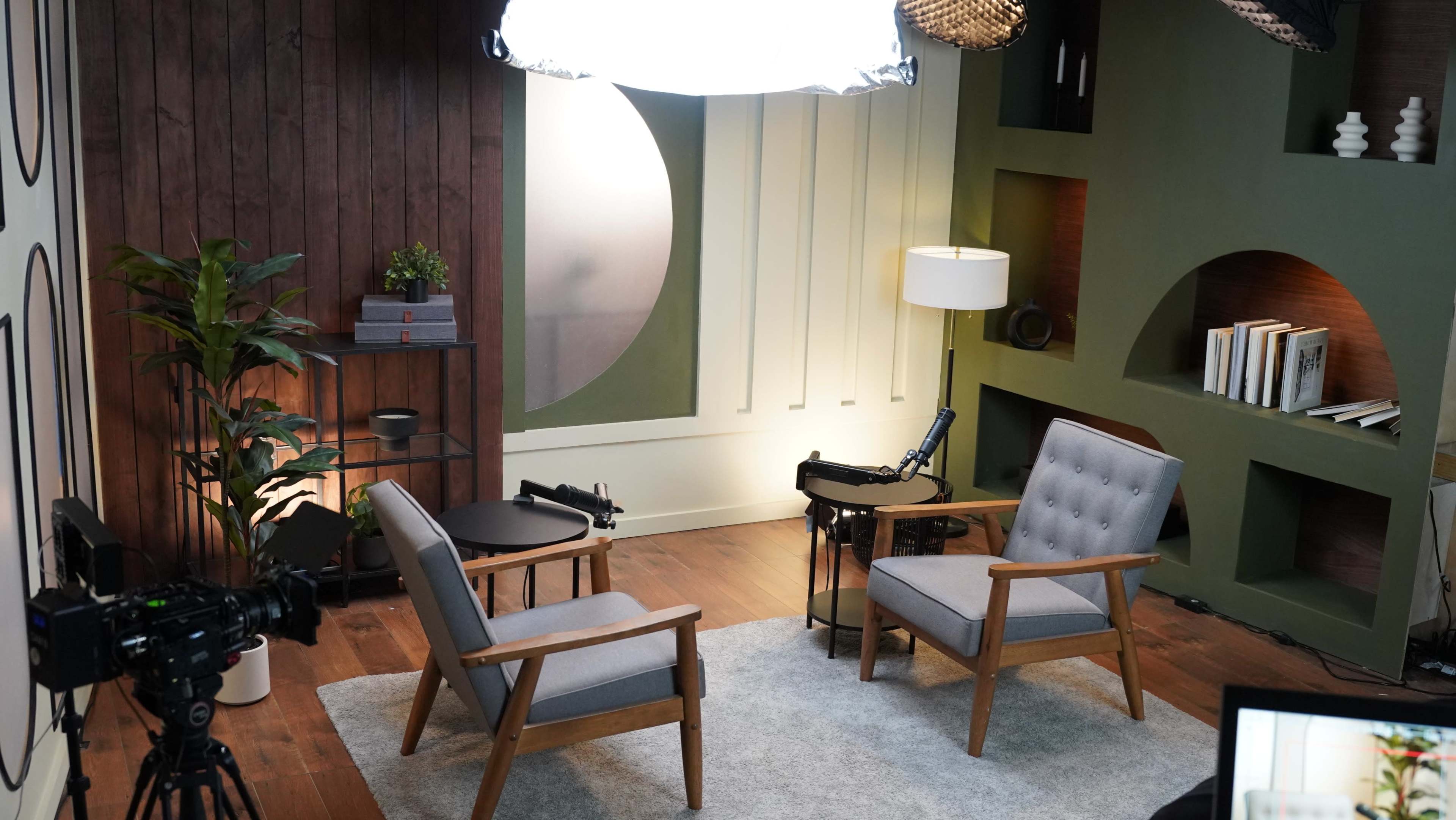The image shows a modern interview set with two gray chairs positioned on a light rug, surrounded by decorative elements such as plants, lamps, and shelves.