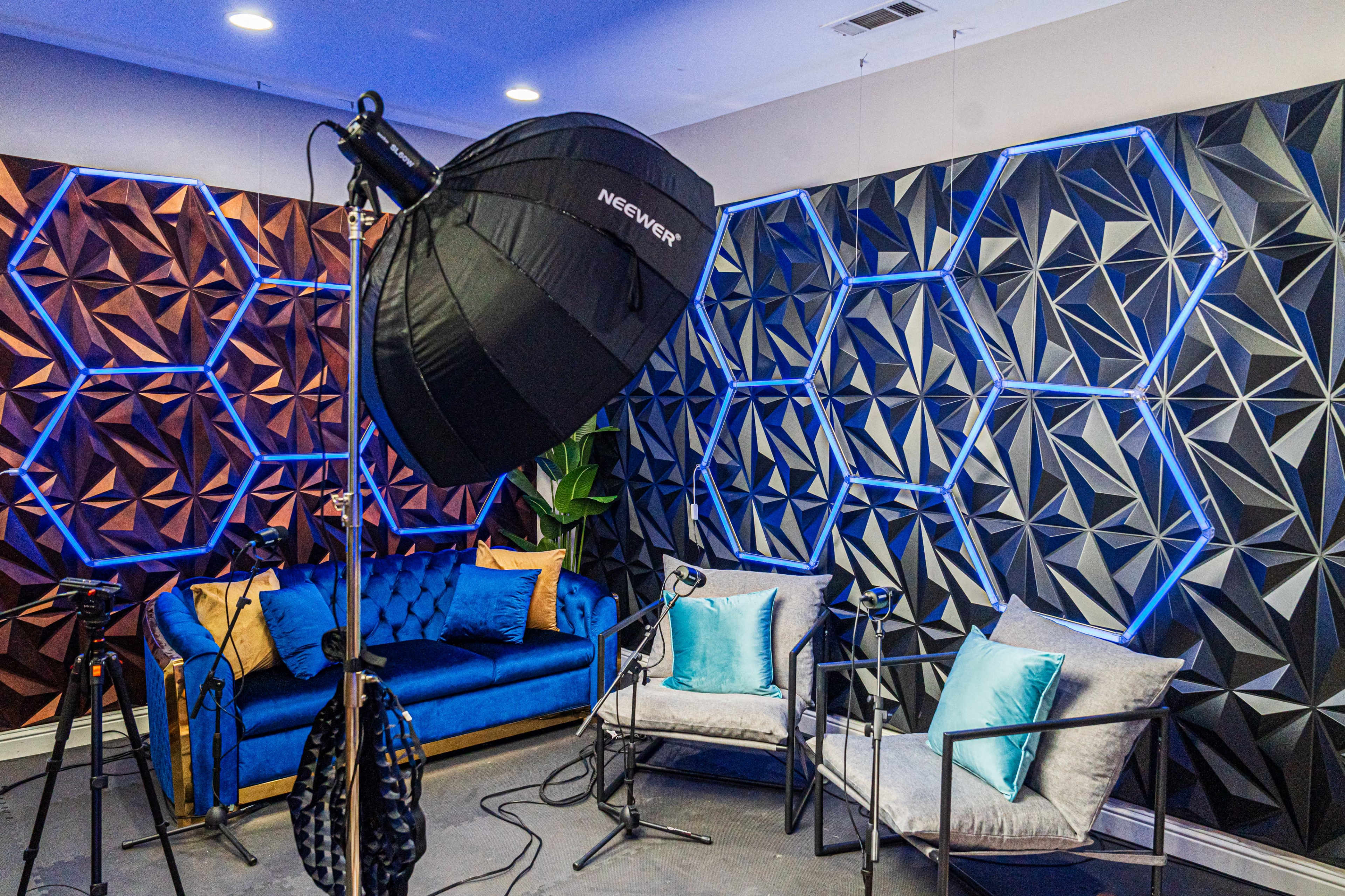 A photography setup features a blue velvet couch and two grey chairs against a backdrop of textured, geometric wall panels illuminated by blue lighting.