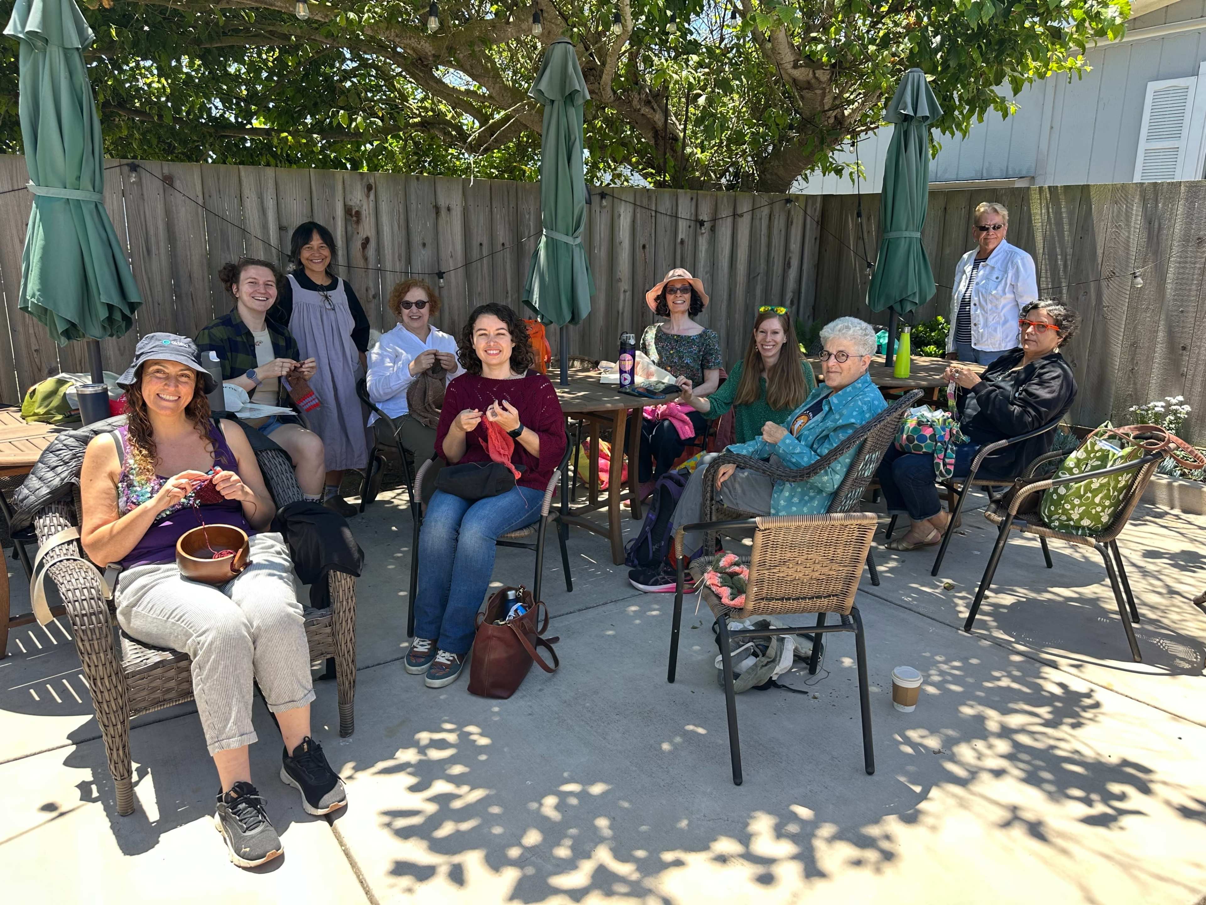 A group of people is gathered outside on a patio, sitting in chairs, some engaged in crafting activities, under green umbrellas and surrounded by trees.