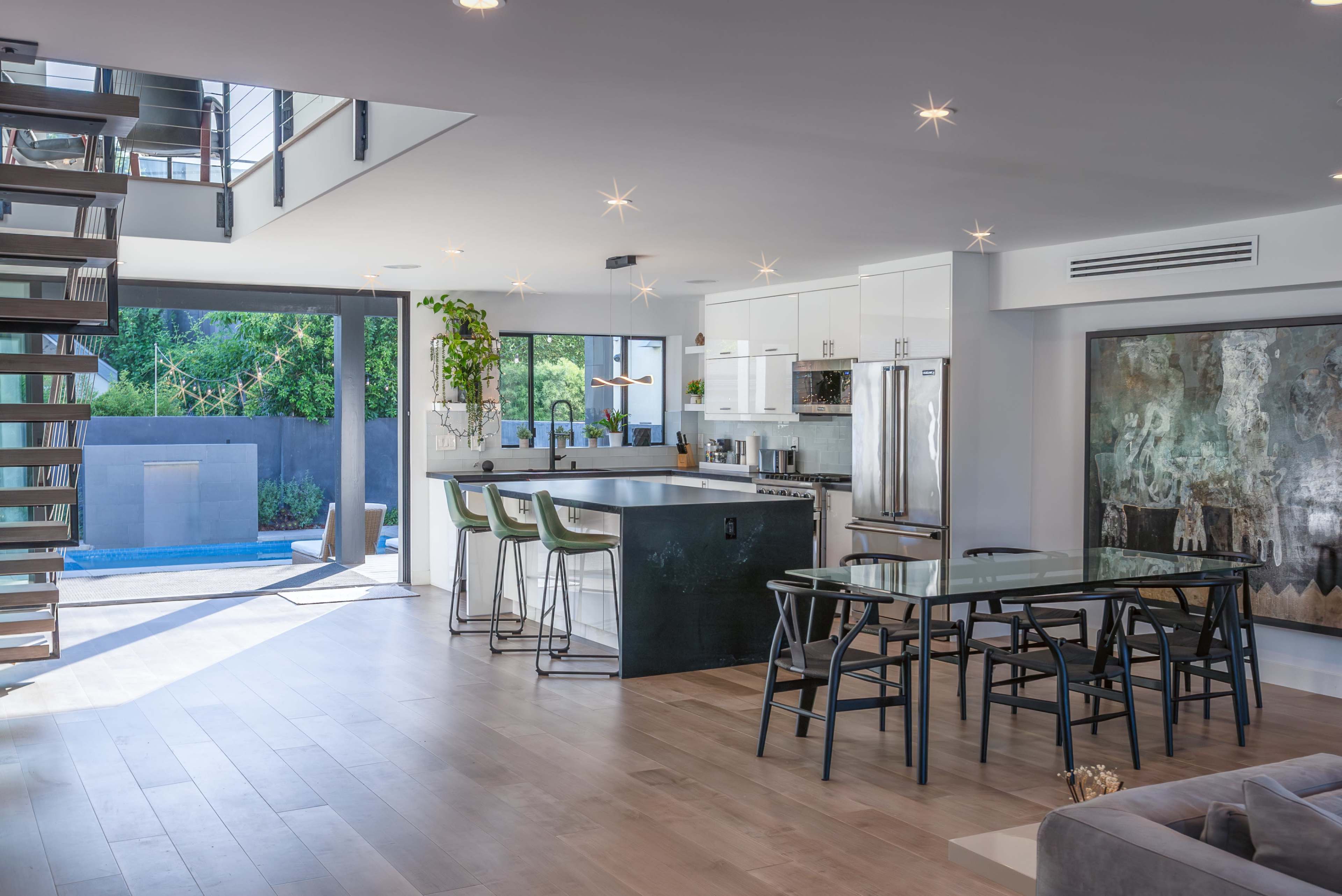 The image shows a modern open-concept kitchen and dining area with a staircase, large windows, and a view of an outdoor pool.