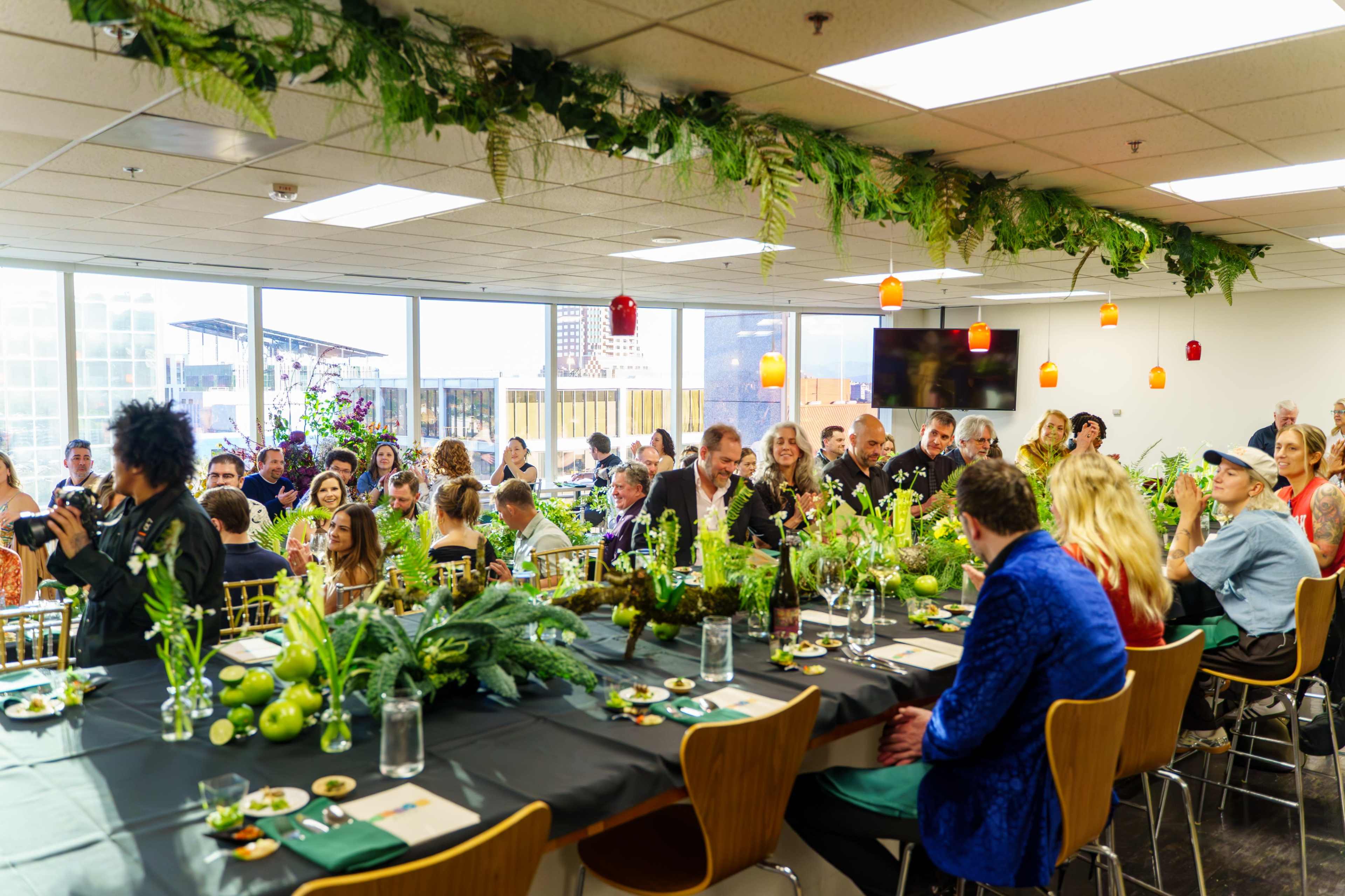 A large gathering of people is seated at a long, decorated table with greenery and food in a well-lit room with large windows.