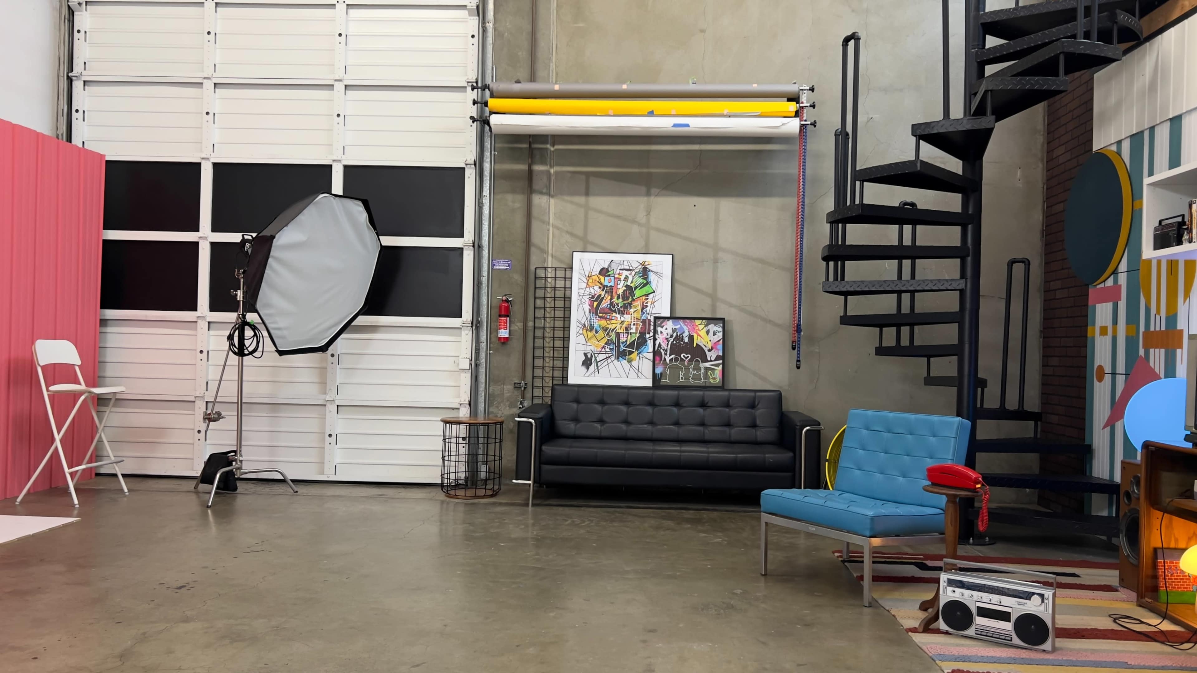 The scene features a spacious studio with a black couch, a blue chair, a spiral staircase, and various artworks on display.