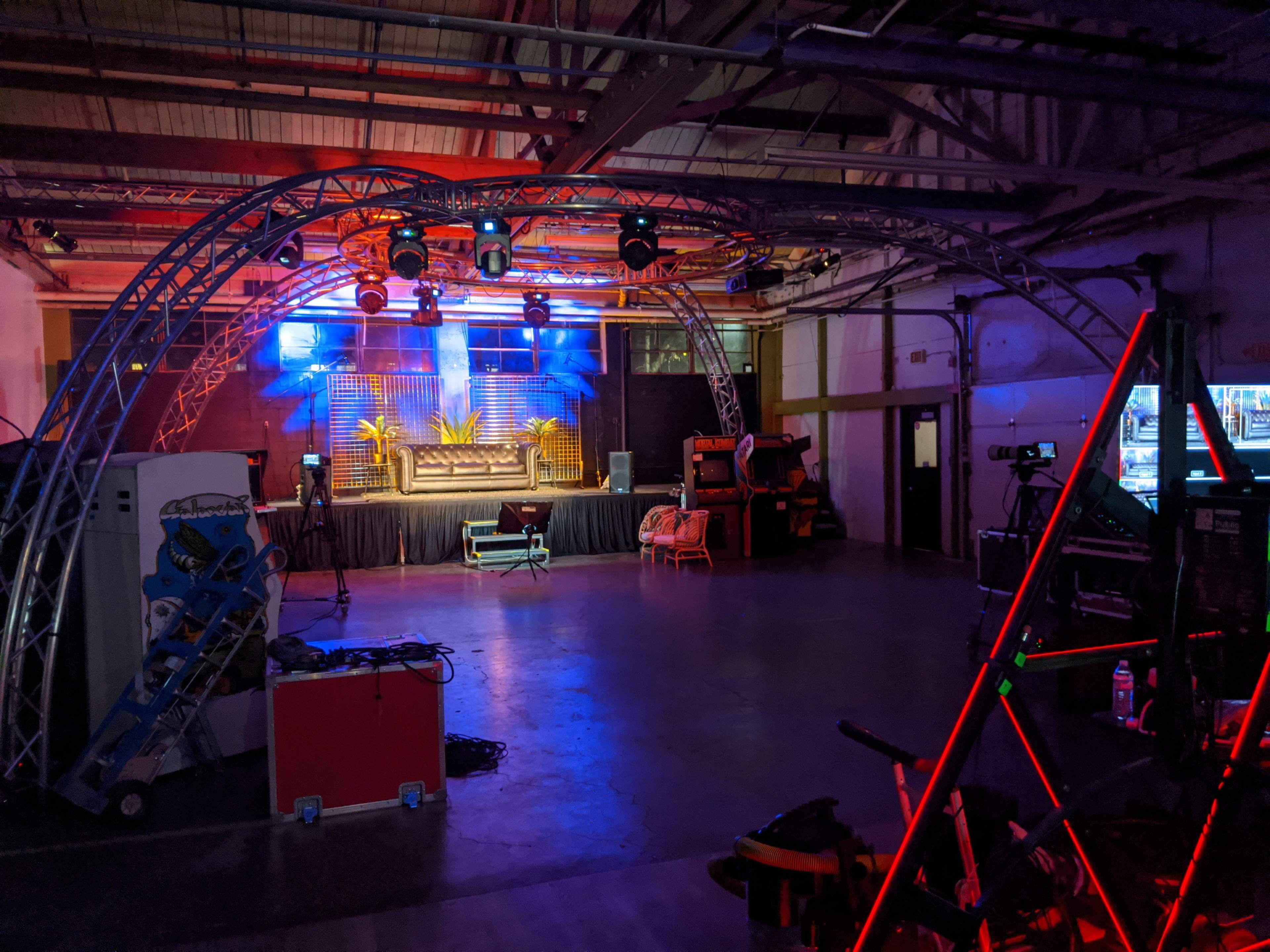 An empty event space with stage lighting, equipment, and a backdrop featuring plants.