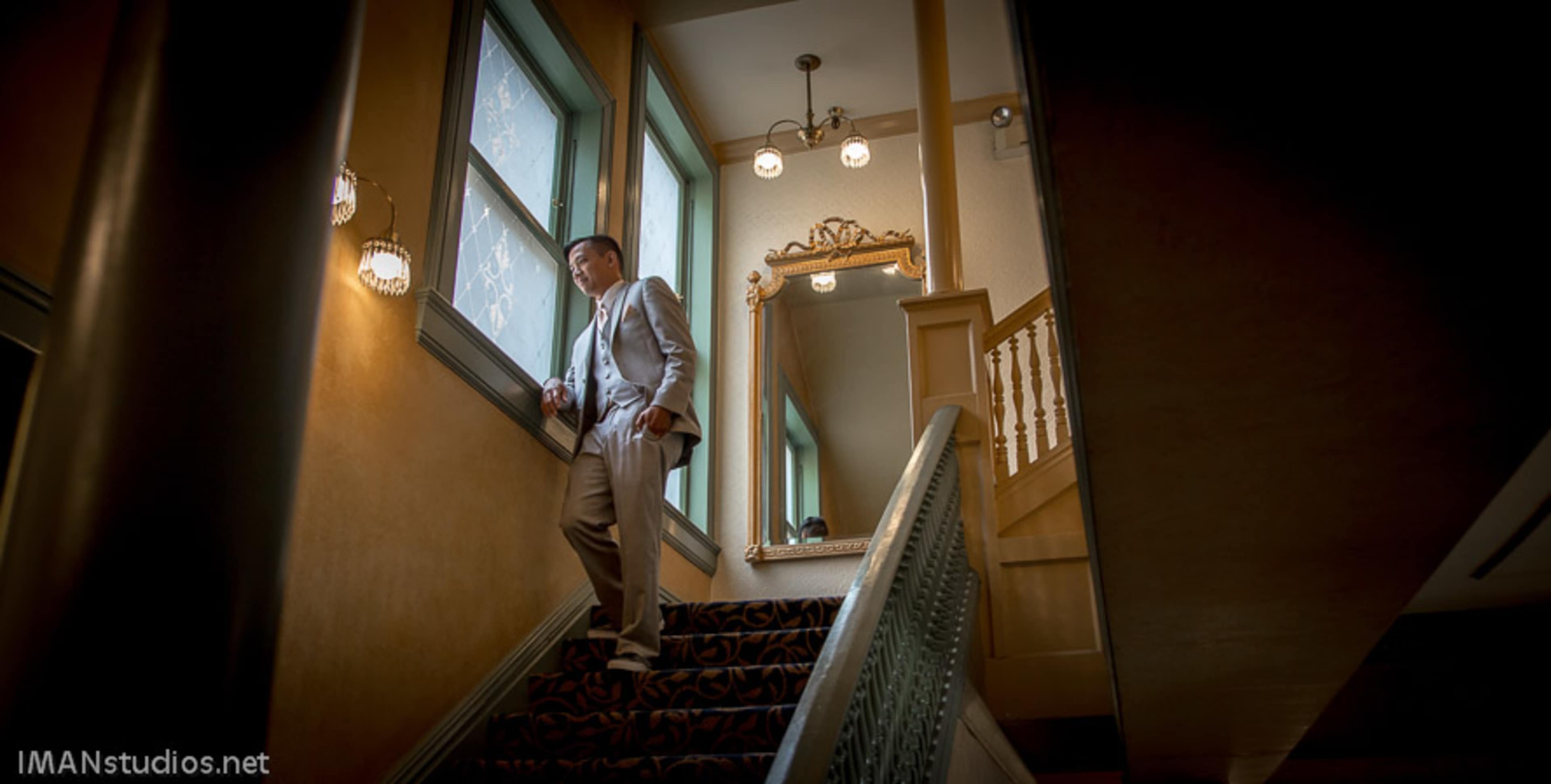 A man in a light suit stands on a staircase next to a window, with a decorative mirror and light fixtures in the background.