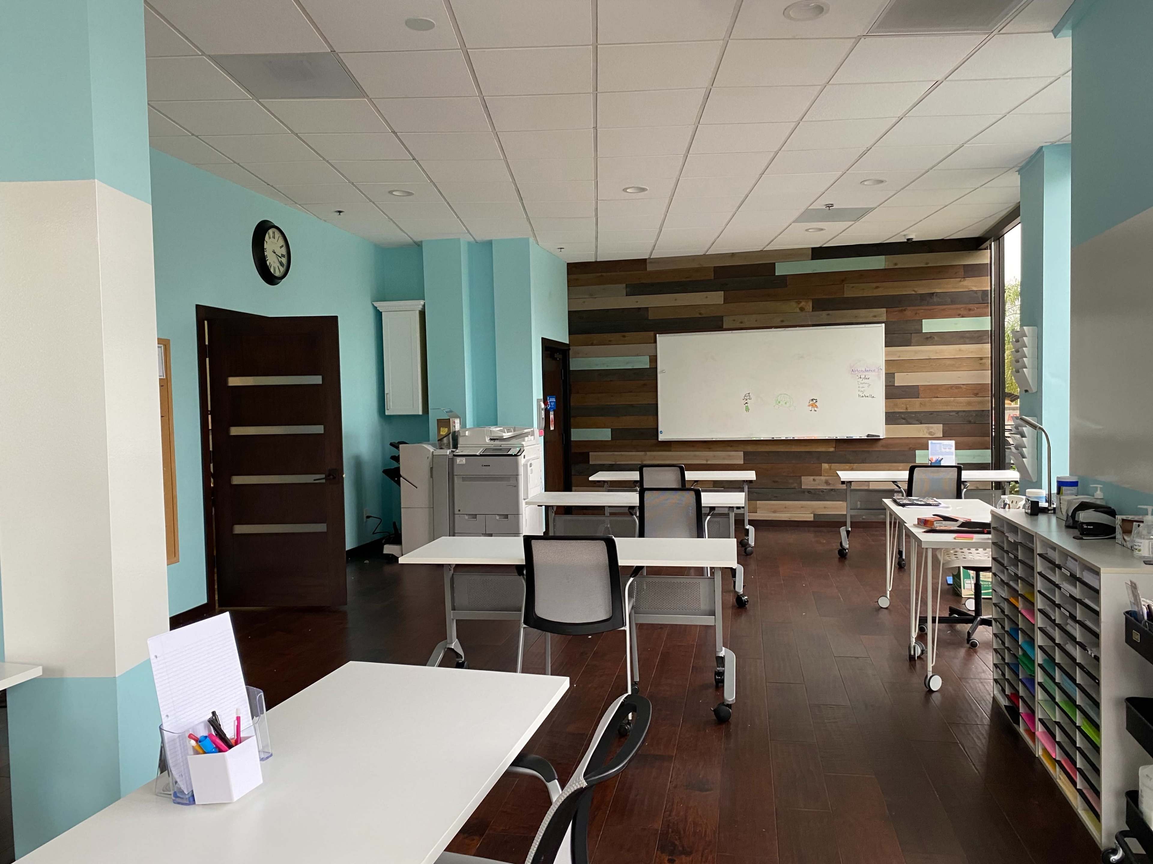A modern classroom with multiple tables arranged for students, a whiteboard on a wooden accent wall, and a clock on the wall.