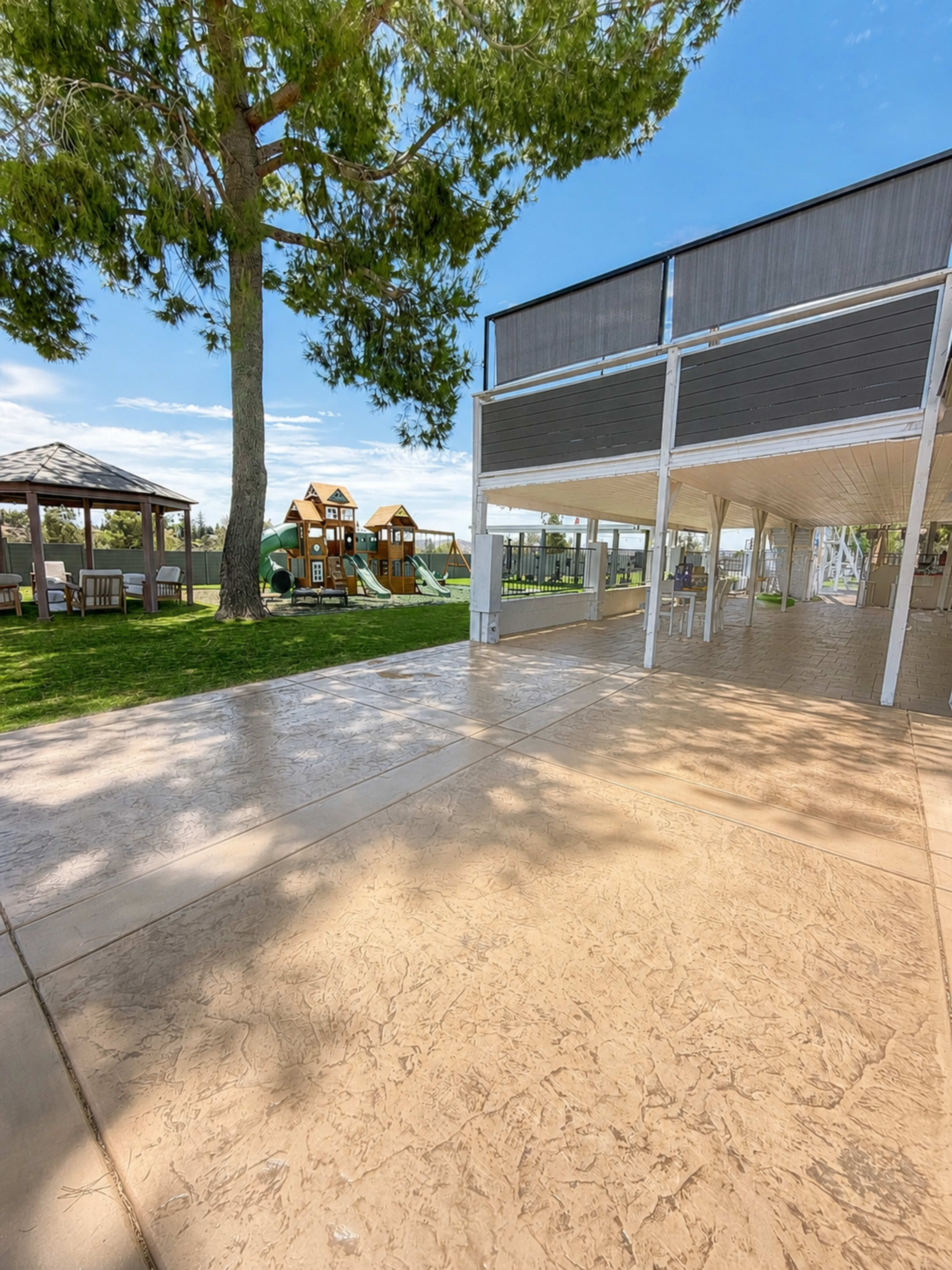 The image shows a landscaped outdoor area with a playground featuring wooden structures, a gazebo, and a spacious concrete patio.