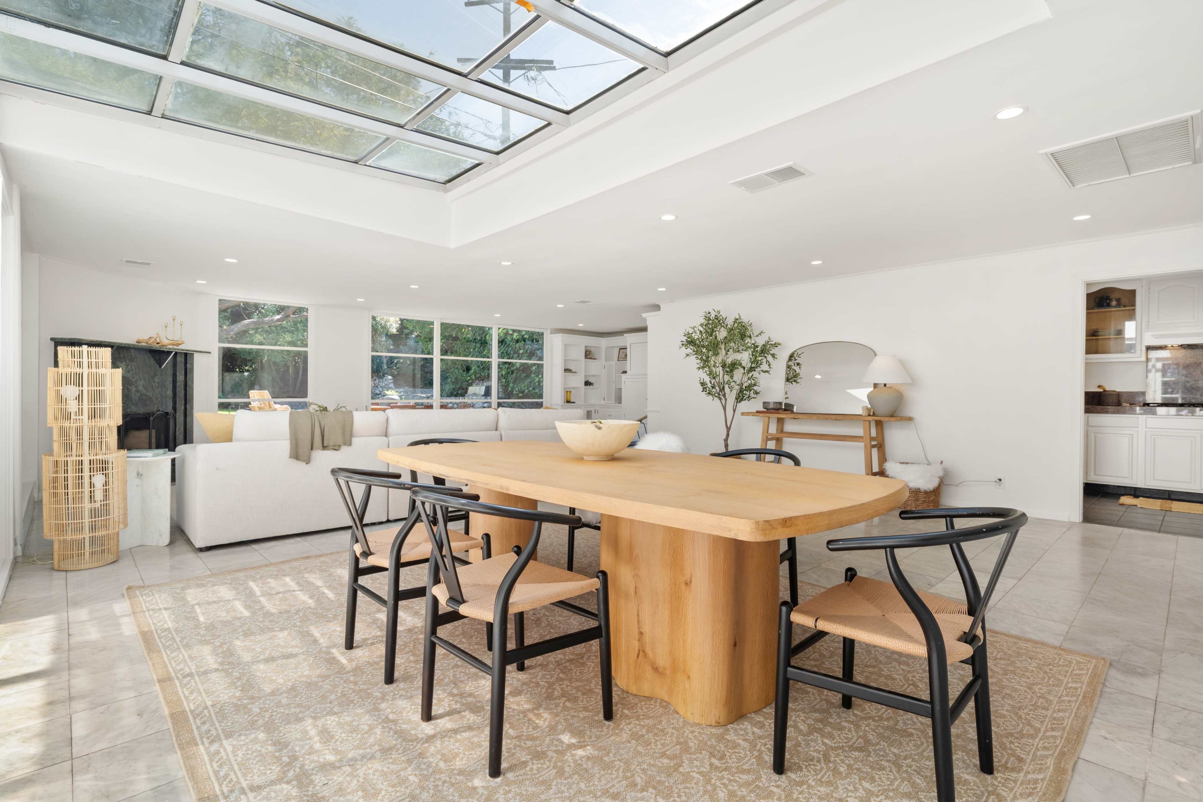 The image shows a modern dining area featuring a large wooden table surrounded by black chairs, with large windows allowing natural light to fill the space.