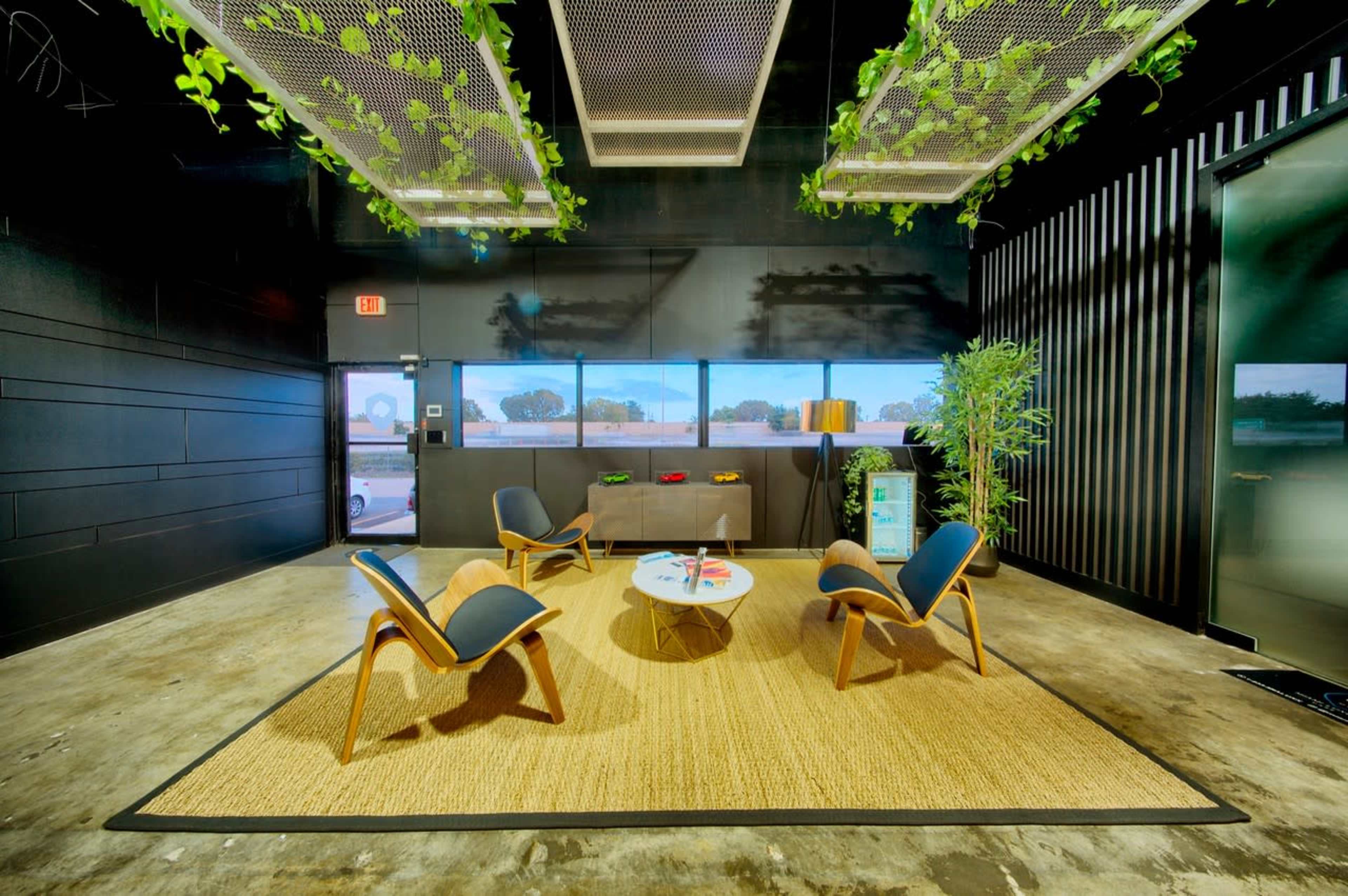 A modern seating area features four chairs around a circular table on a woven rug, with large windows displaying an outdoor view and greenery embellishing the ceiling.