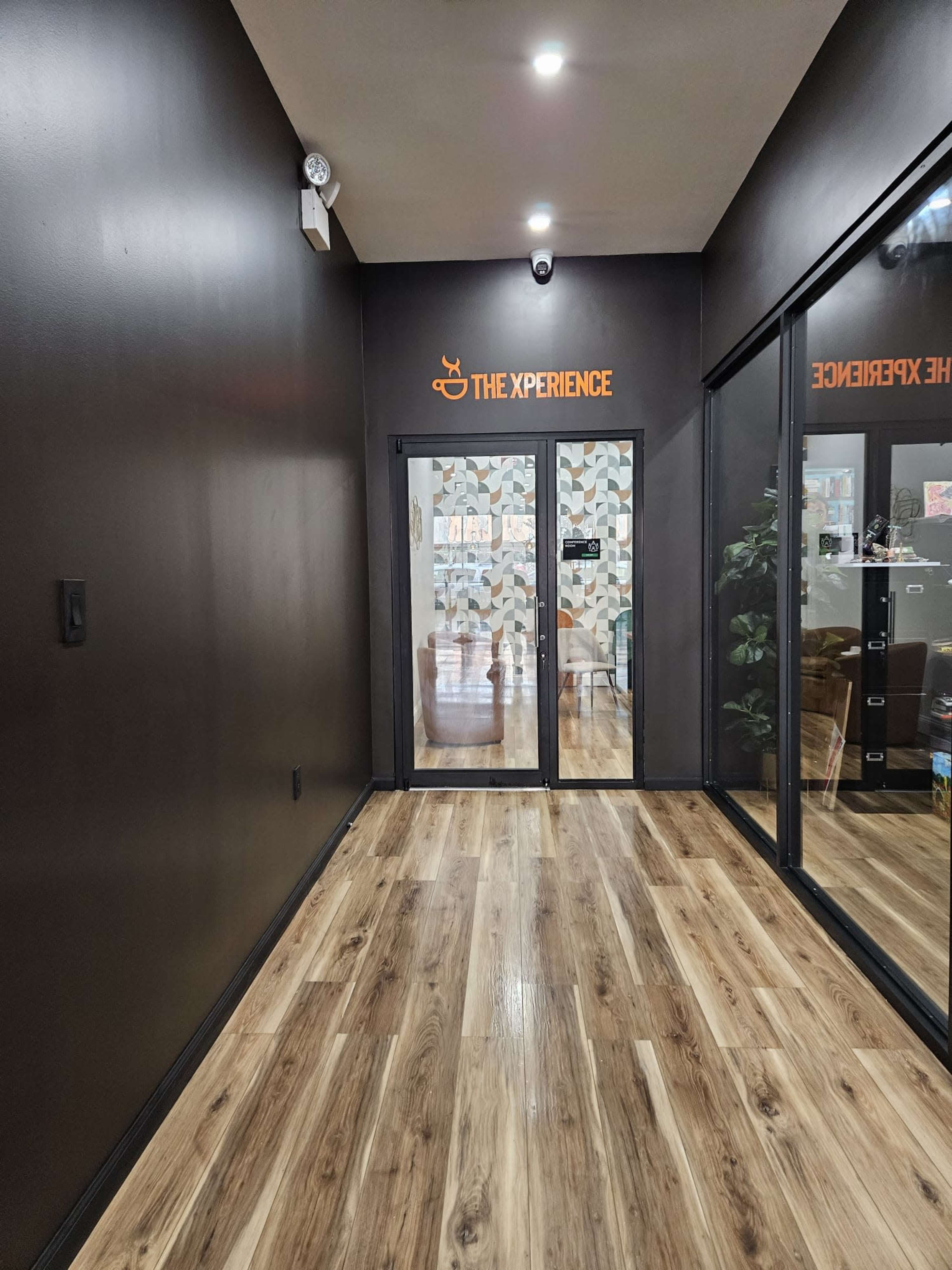 A dimly lit hallway with dark walls, wooden flooring, and glass double doors leading to a brightly decorated room.