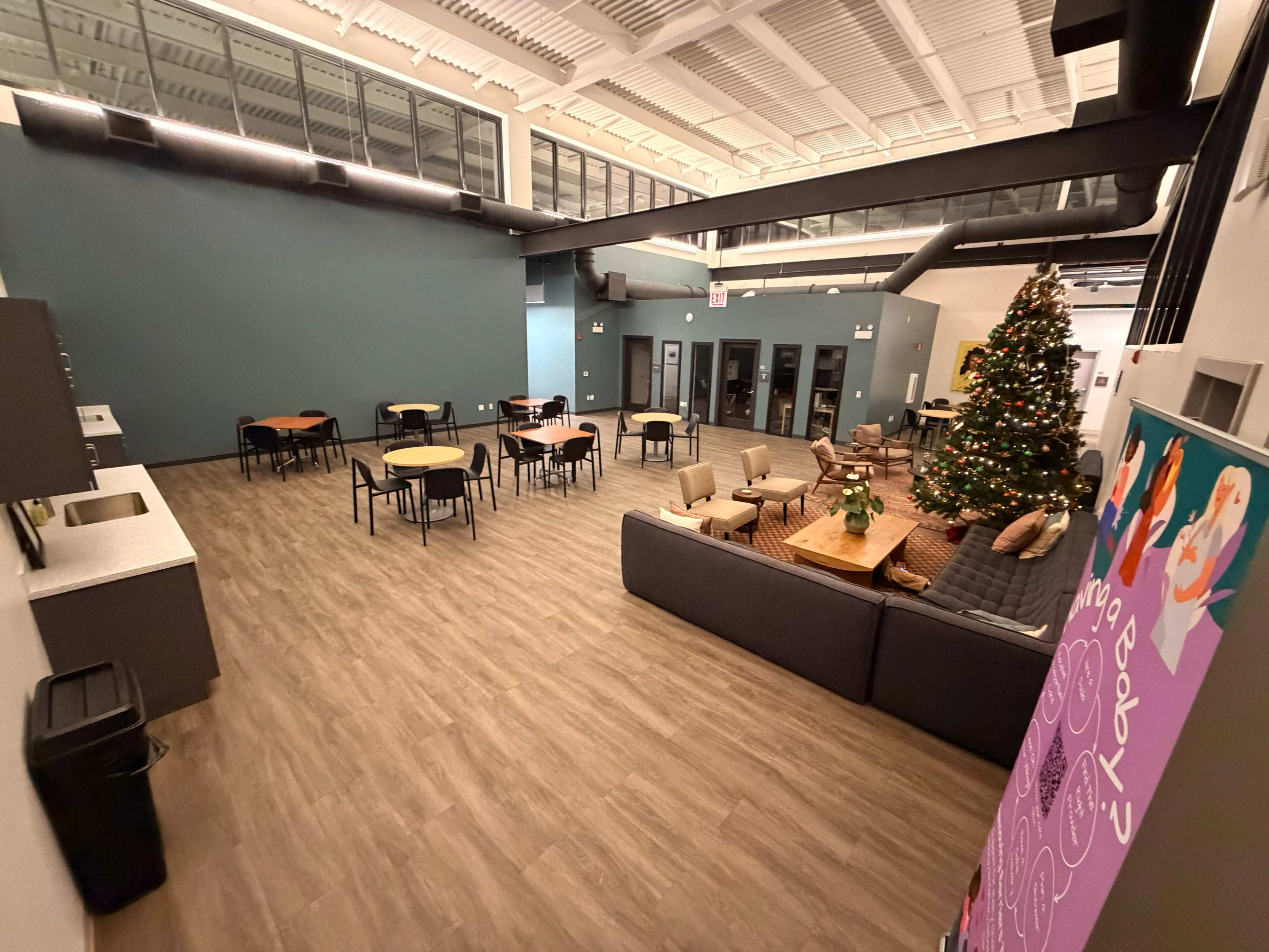 The image shows a large, spacious common area with tables, chairs, a sofa, and a Christmas tree decorated with lights and ornaments.