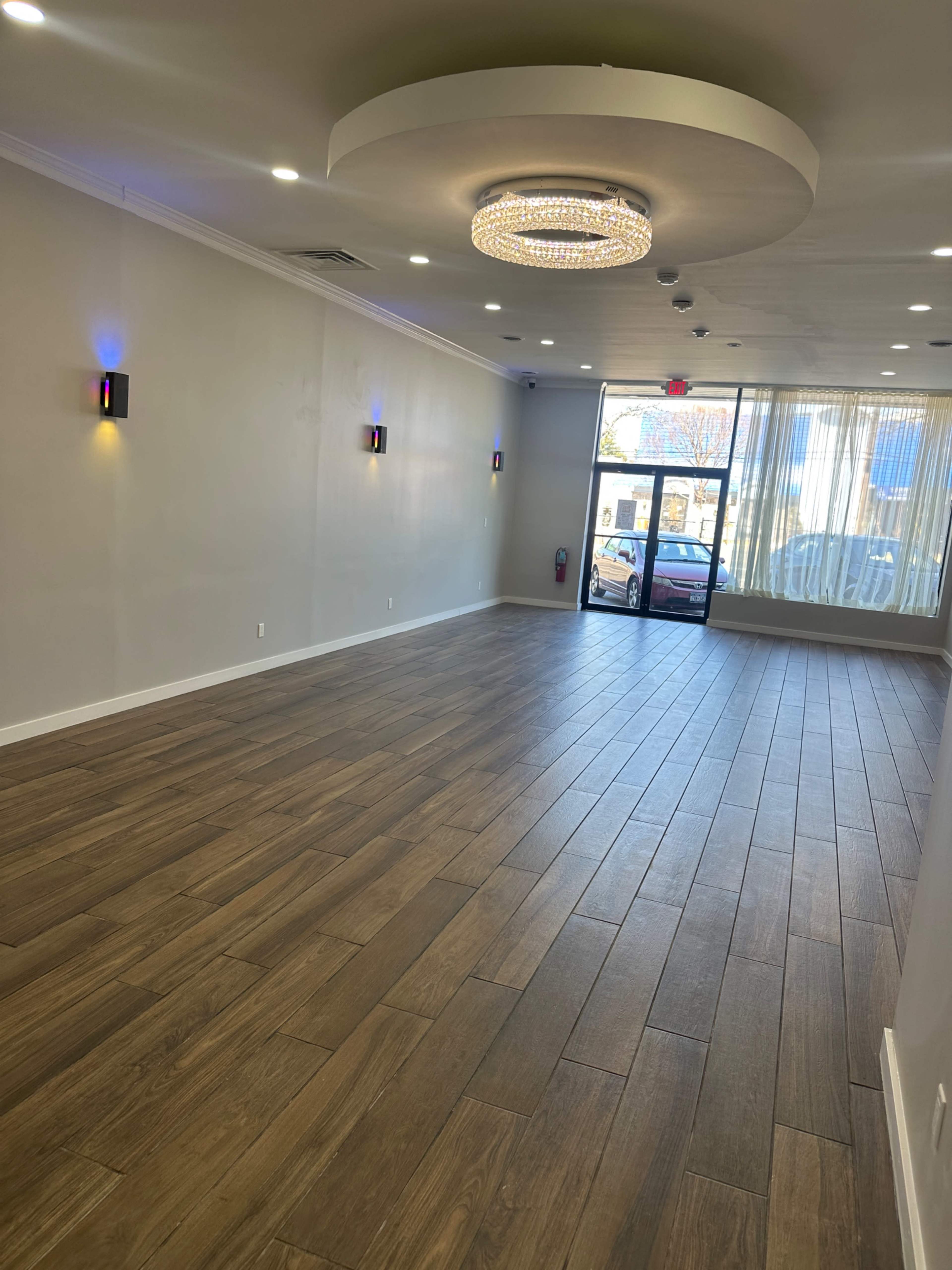 The image shows an empty interior space with wooden flooring, a round chandelier on the ceiling, and large windows at the entrance.