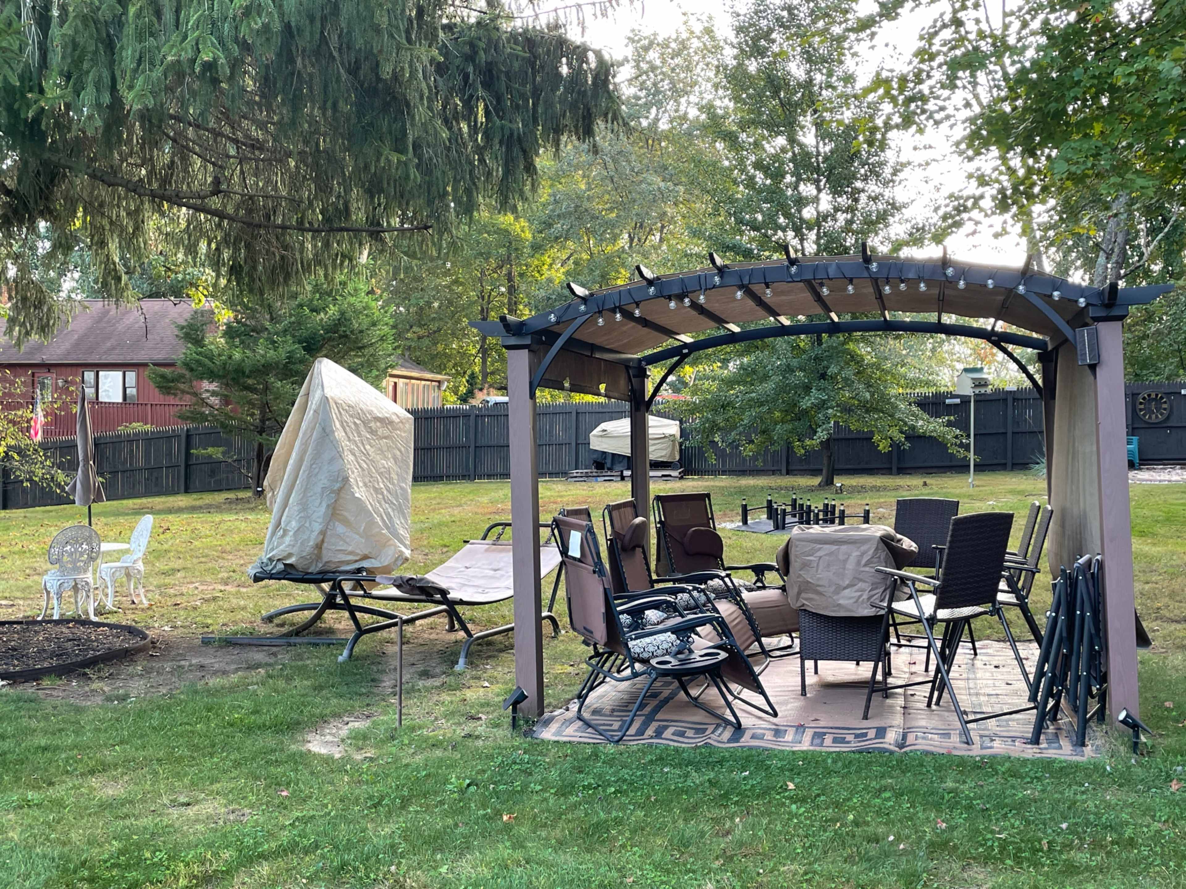 A backyard patio features a covered seating area with a table and chairs surrounded by trees and a grassy lawn.