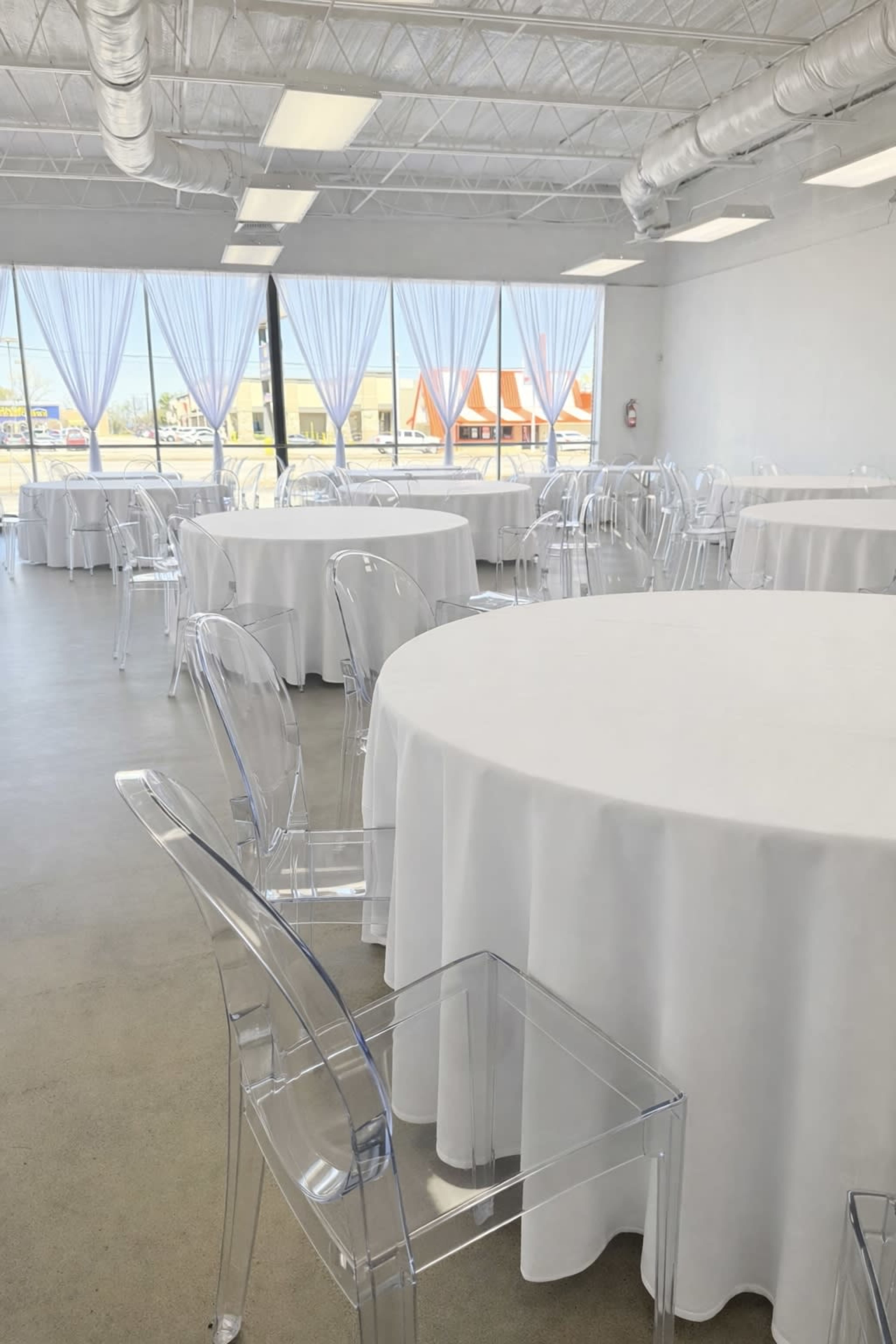 The image shows a spacious event room with several round tables covered in white tablecloths and clear acrylic chairs, illuminated by natural light from large windows.