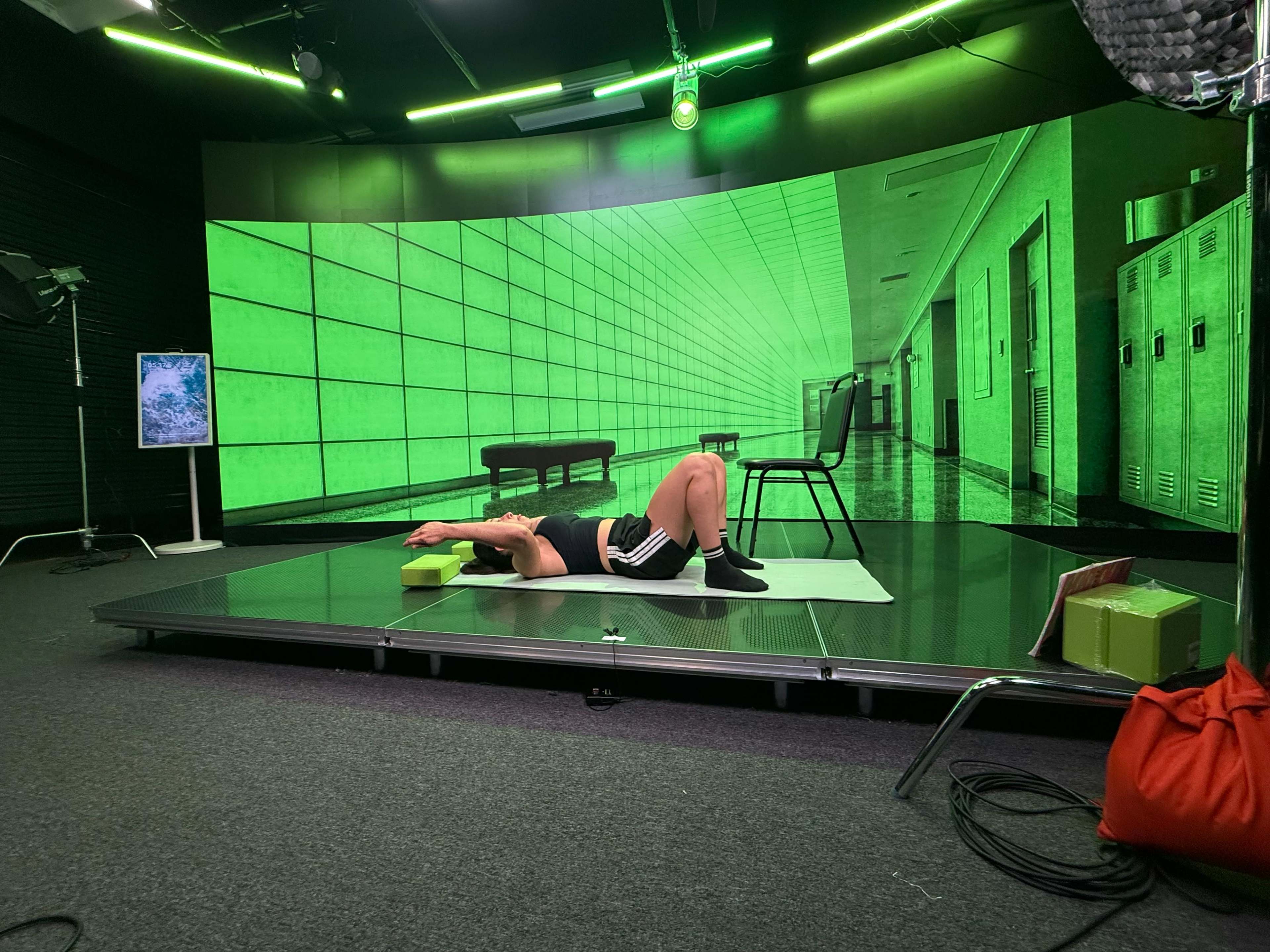 A person lies on a yoga mat in a studio setting with a green backdrop featuring a corridor and lockers.