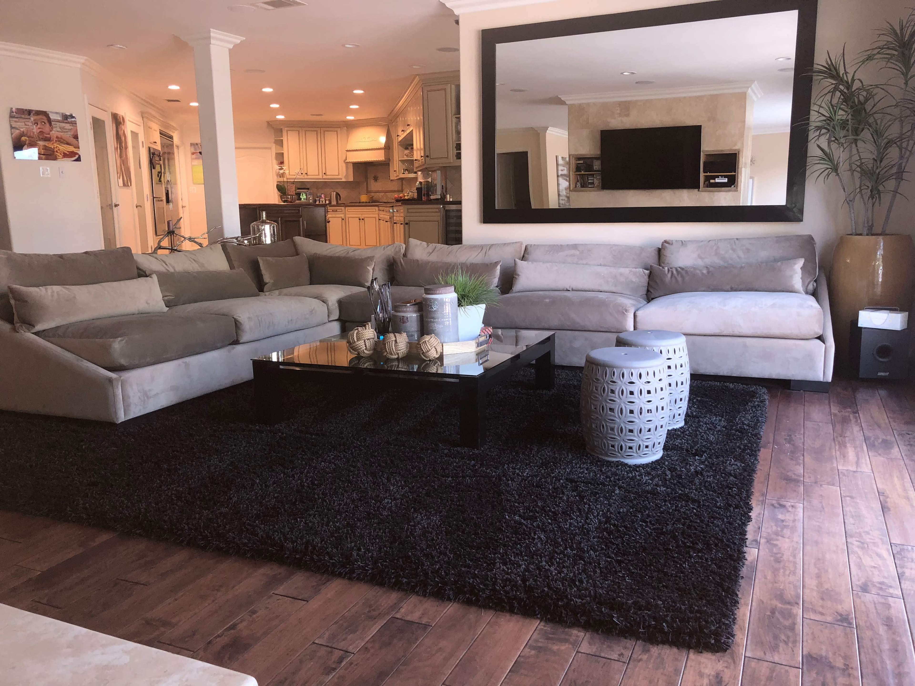 The image shows a modern living room featuring a large gray sectional sofa, a black coffee table on a textured rug, and an open kitchen in the background.