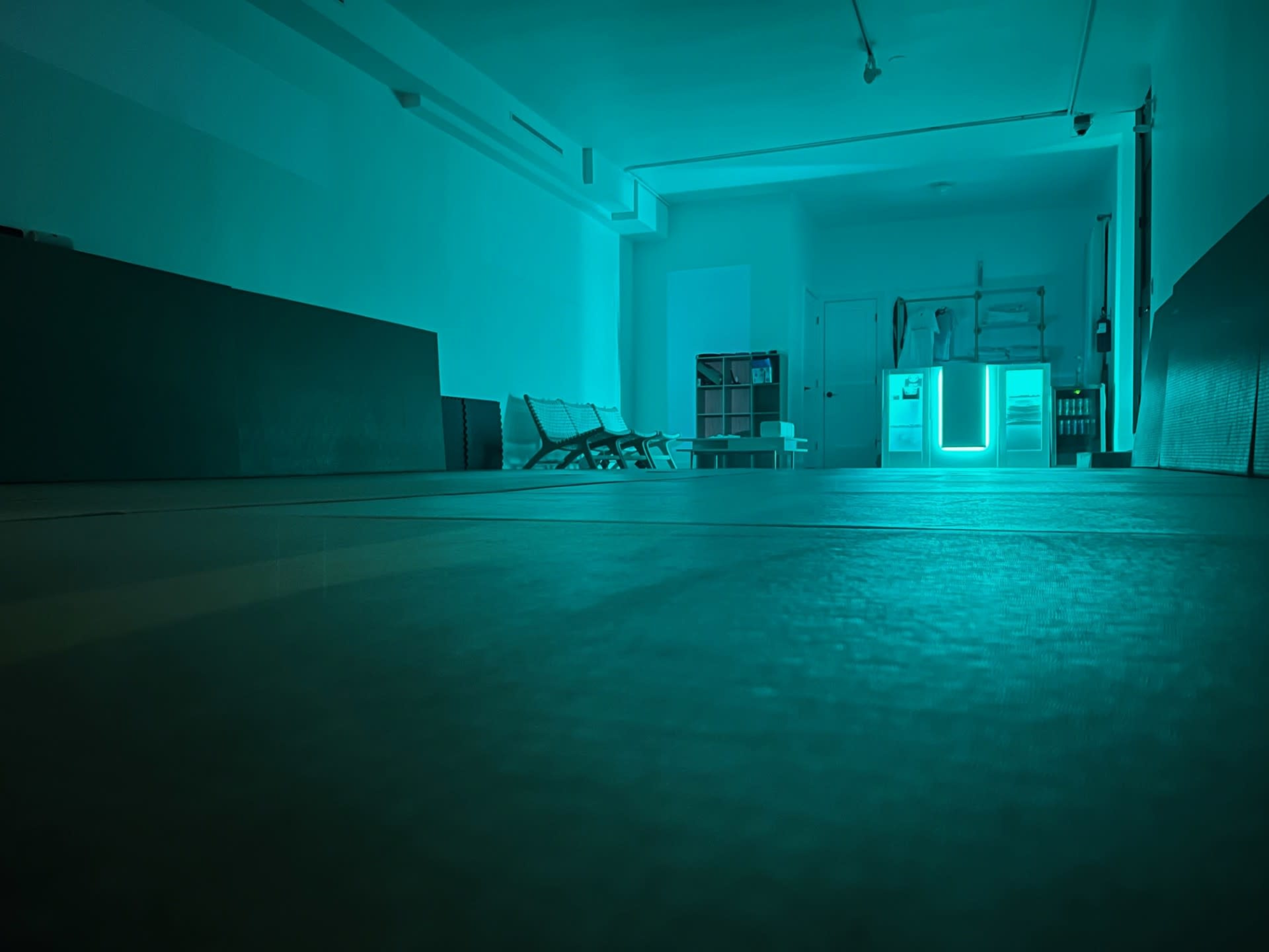 The image shows a dimly lit, empty room with a reflective floor, featuring chairs along one side and a glowing door in the background.