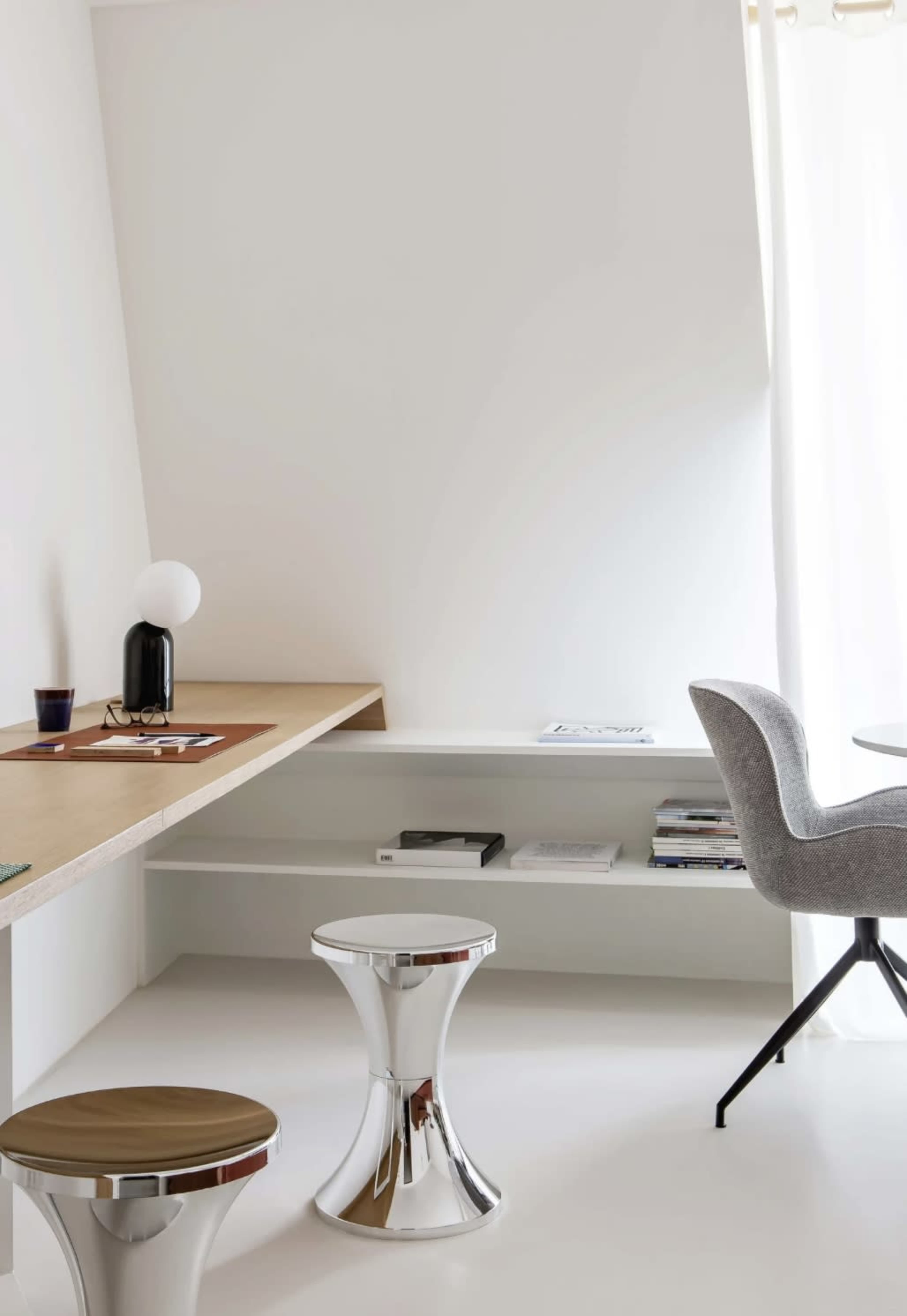 The image shows a minimalist room with a wooden desk, two metallic stools, a gray upholstered chair, and a bookshelf against a white wall.