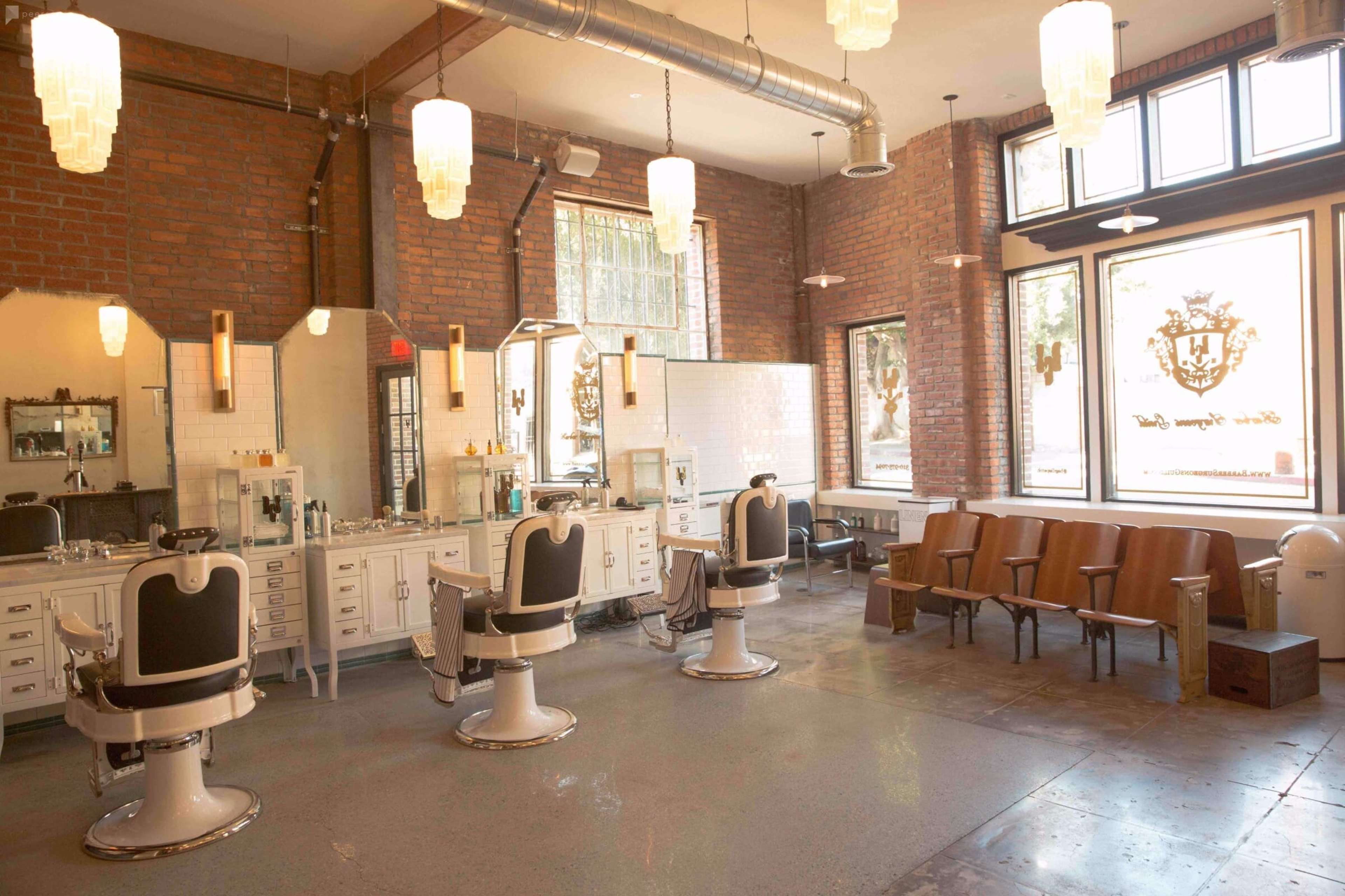 A barber shop interior featuring three vintage barber chairs, large mirrors, and wooden seating under bright light fixtures in a brick-walled setting.
