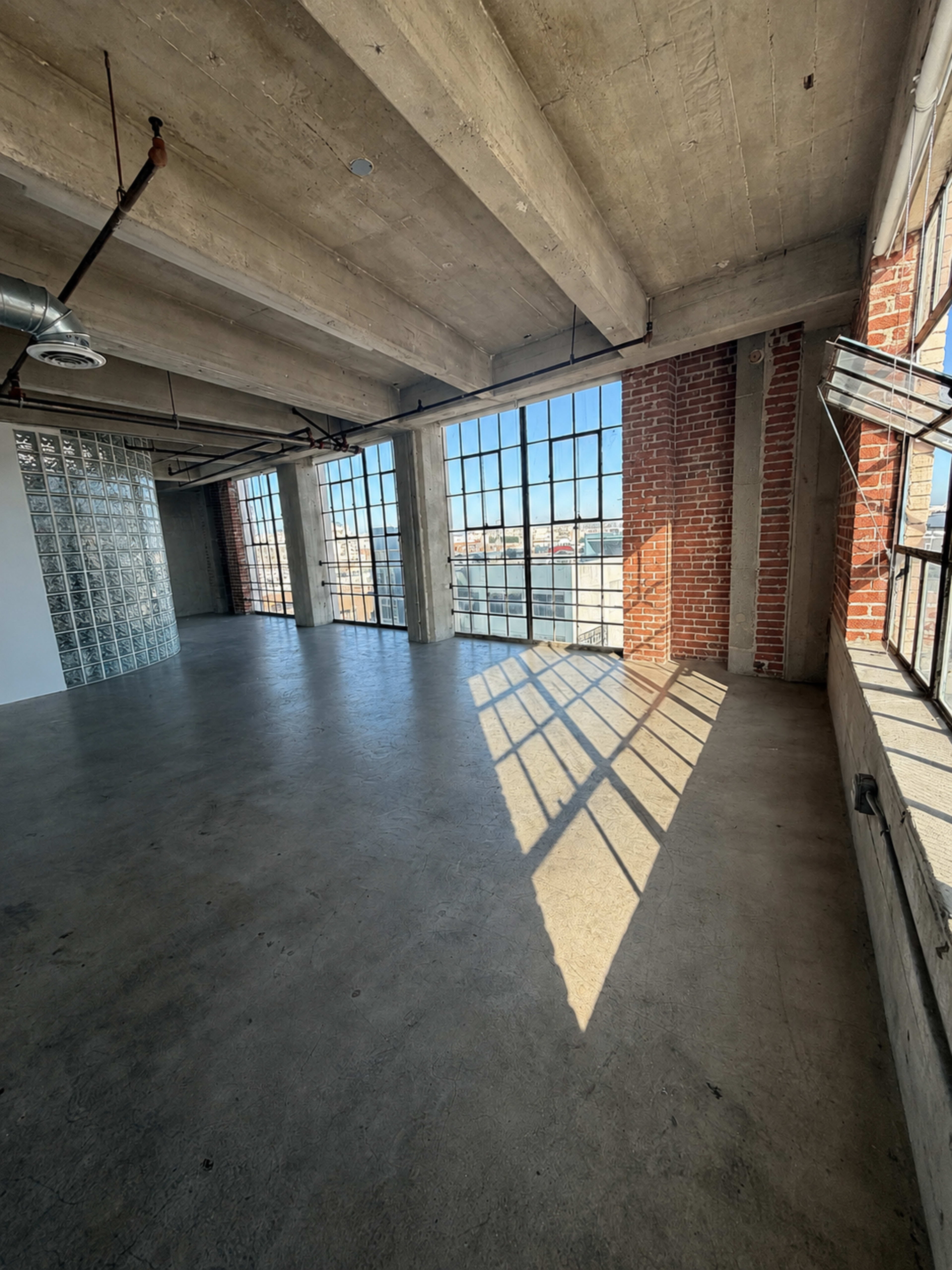 The image shows a spacious industrial-style room with large windows, concrete flooring, exposed brick walls, and beams overhead.