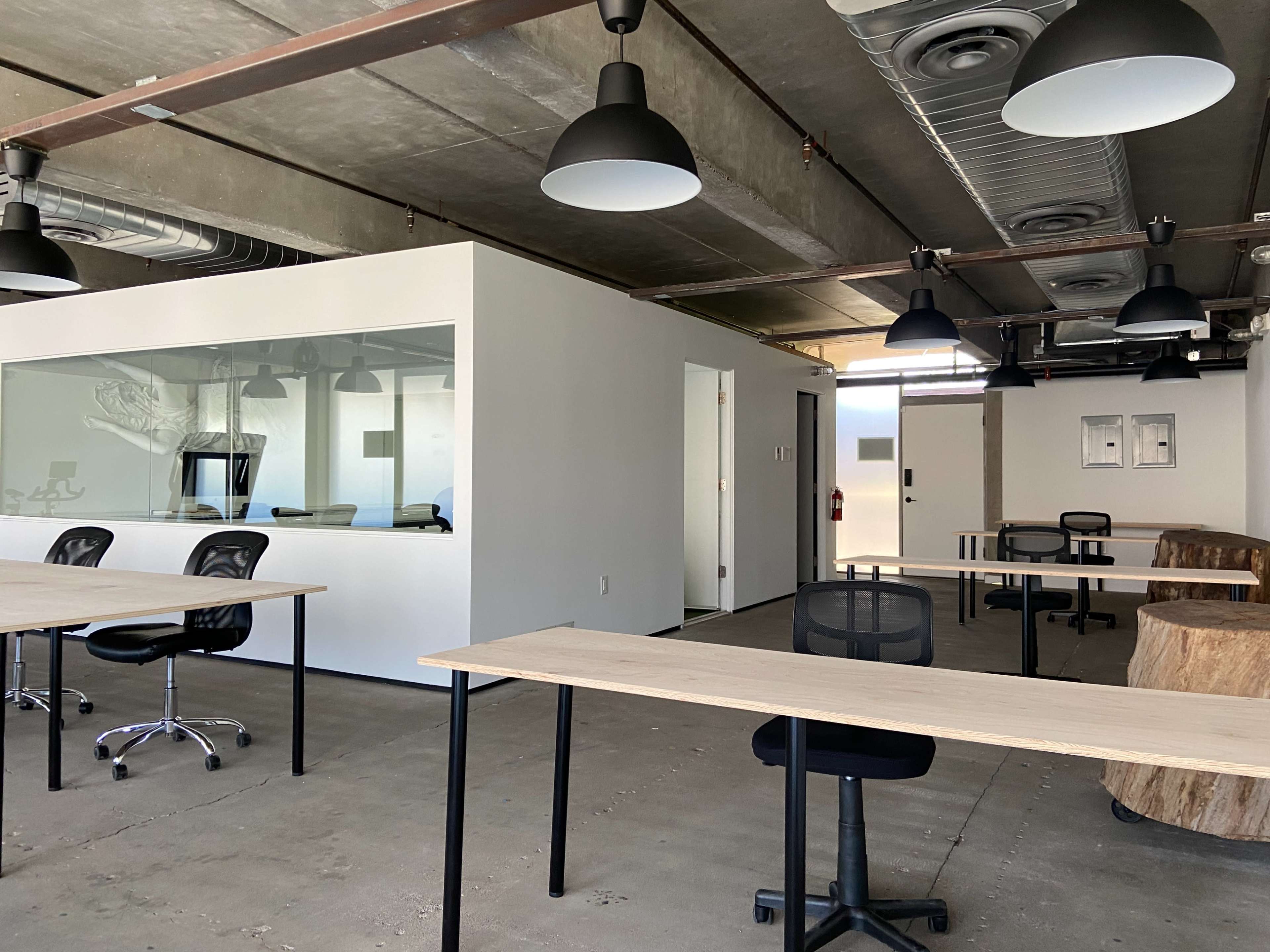 A modern office space with bare concrete floors, several desks with chairs, and a glass-walled meeting room.