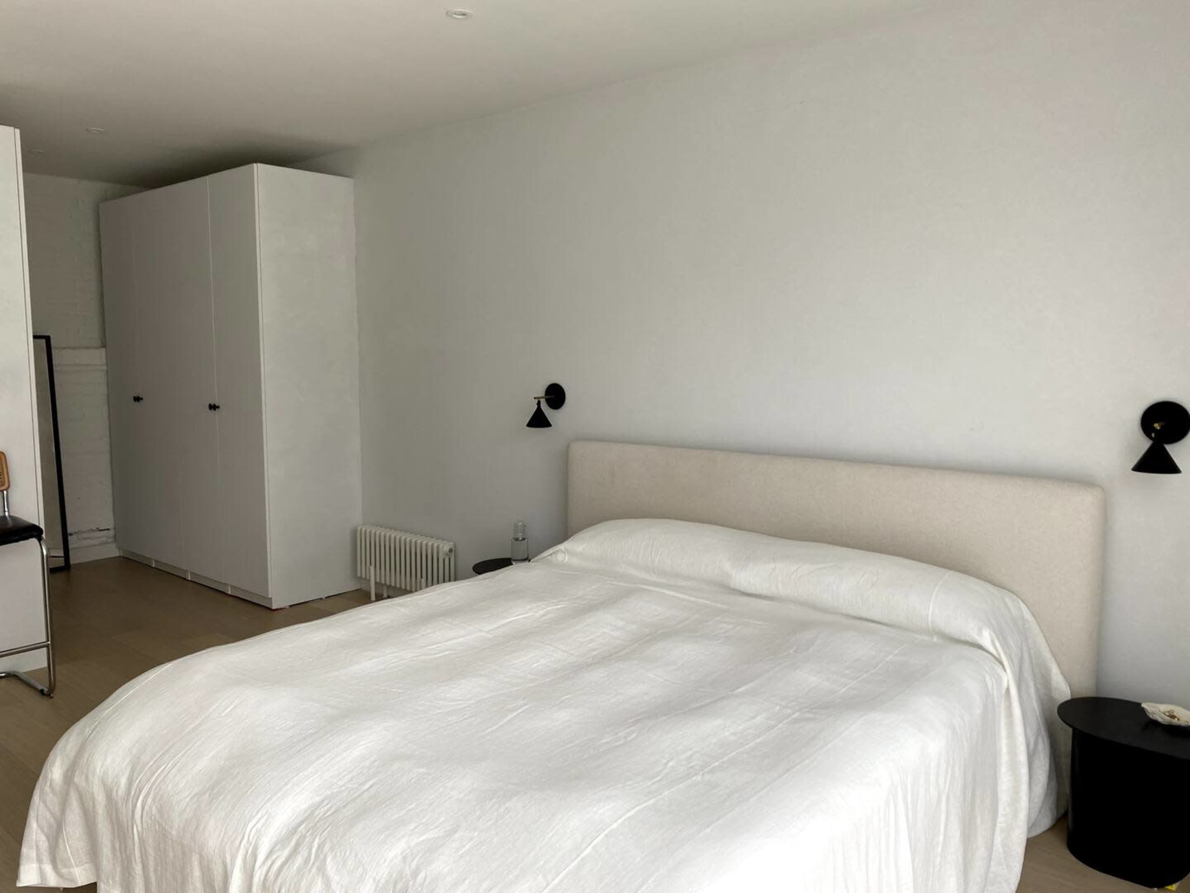 A minimalistic bedroom with a white bedspread, a headboard, two wall-mounted lamps, and a wardrobe.