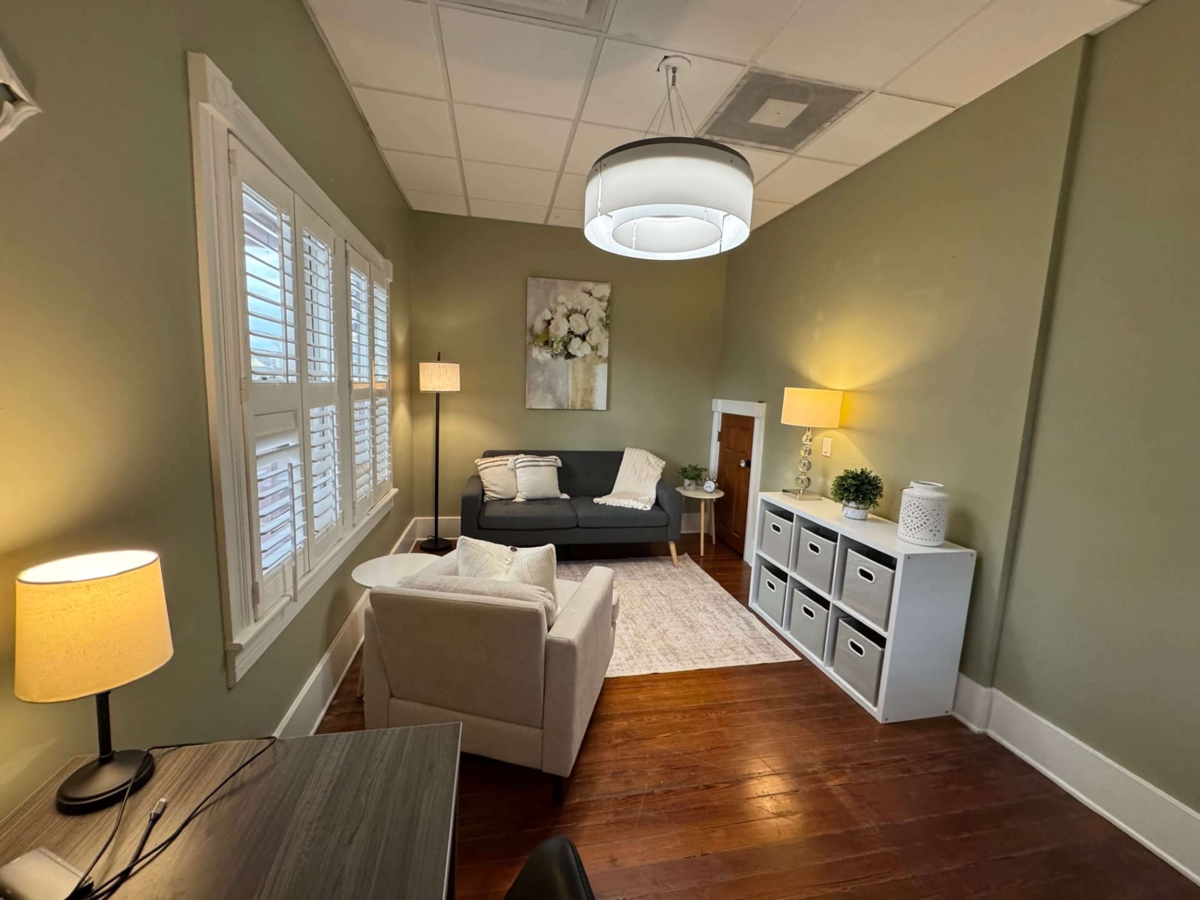 The image shows a small, well-lit room with a gray sofa, a white armchair, a desk, and decorative elements including lamps and storage boxes.
