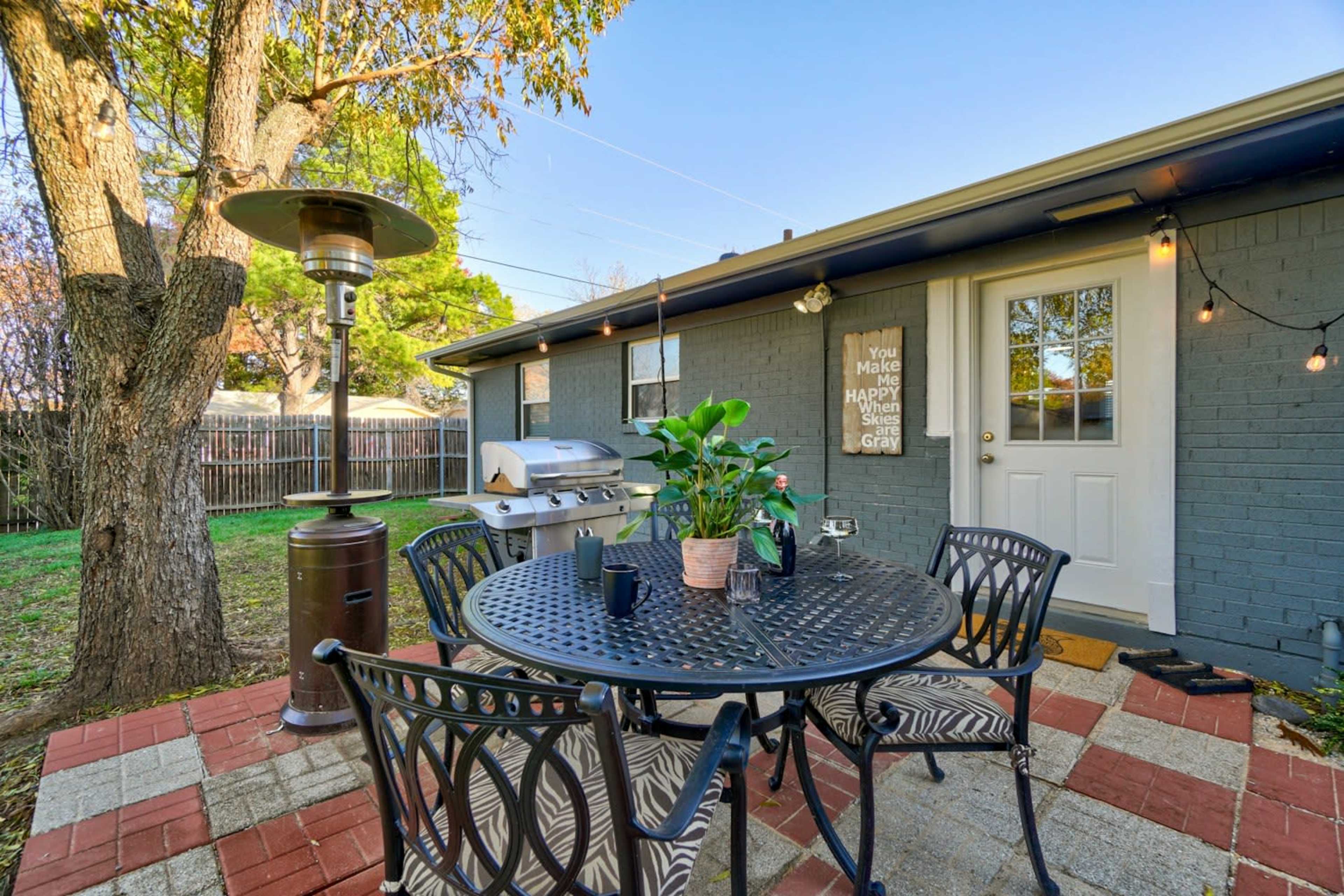 A patio with a round table, chairs, and a grill is set against a house with a door and string lights, surrounded by greenery.