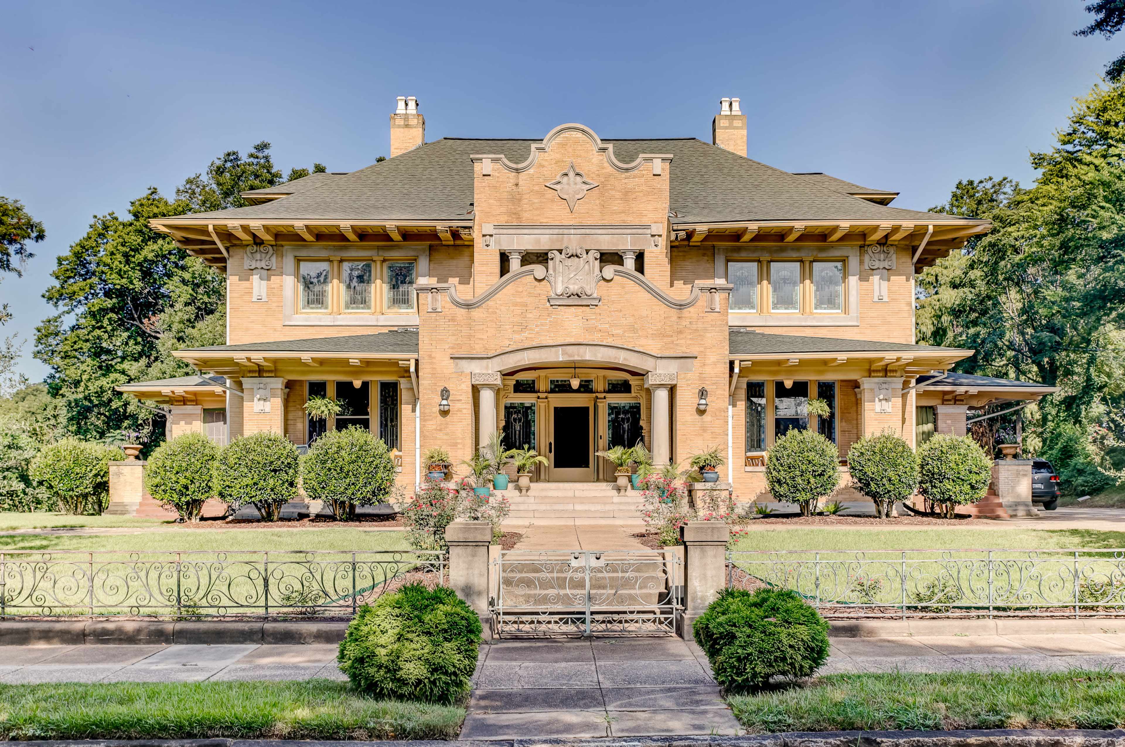 A large, two-story brick house with a decorative facade, surrounded by manicured shrubs and a wrought-iron fence.