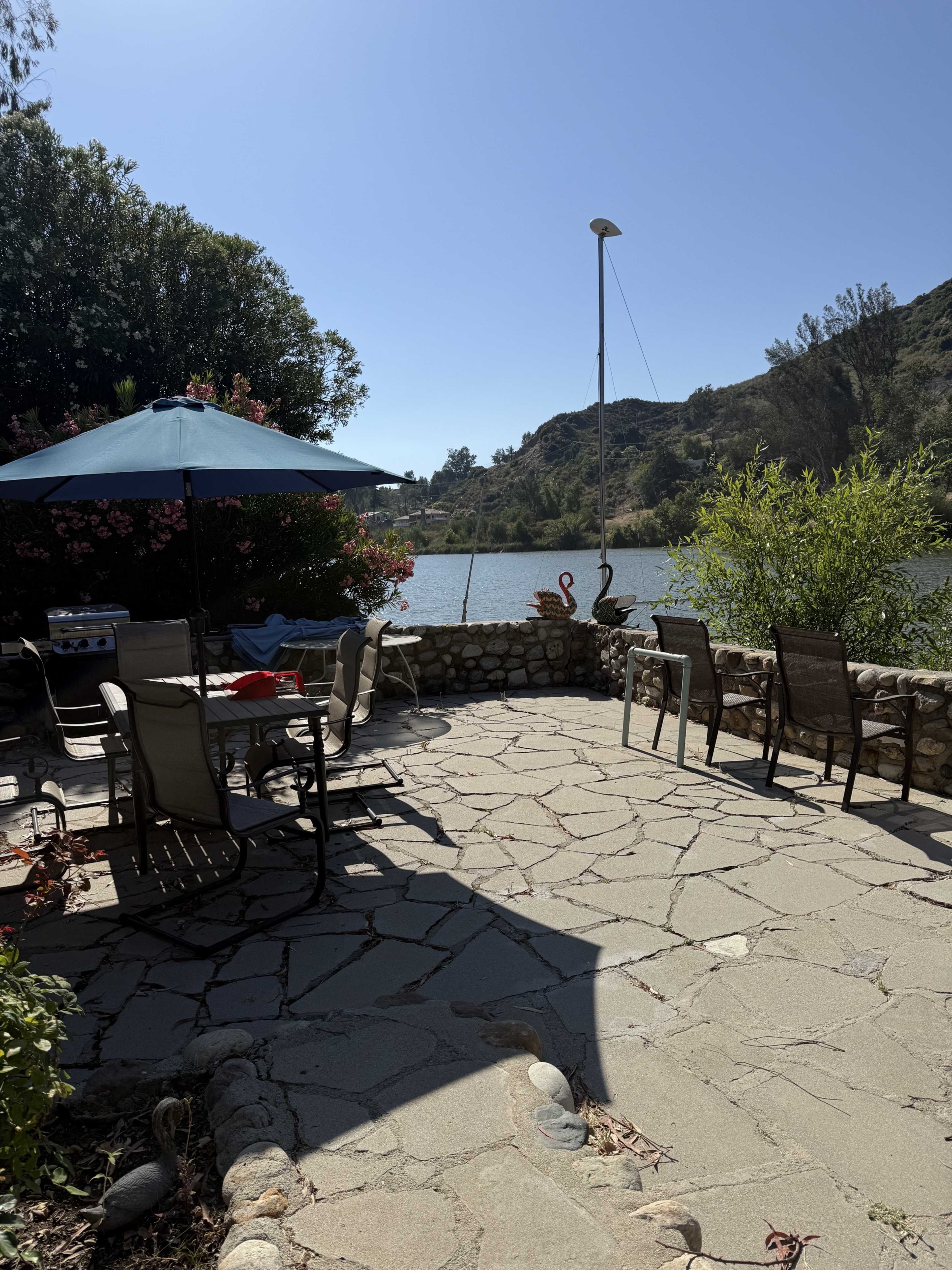 A stone-paved patio near a body of water features several outdoor chairs and a large umbrella, surrounded by greenery and hills.
