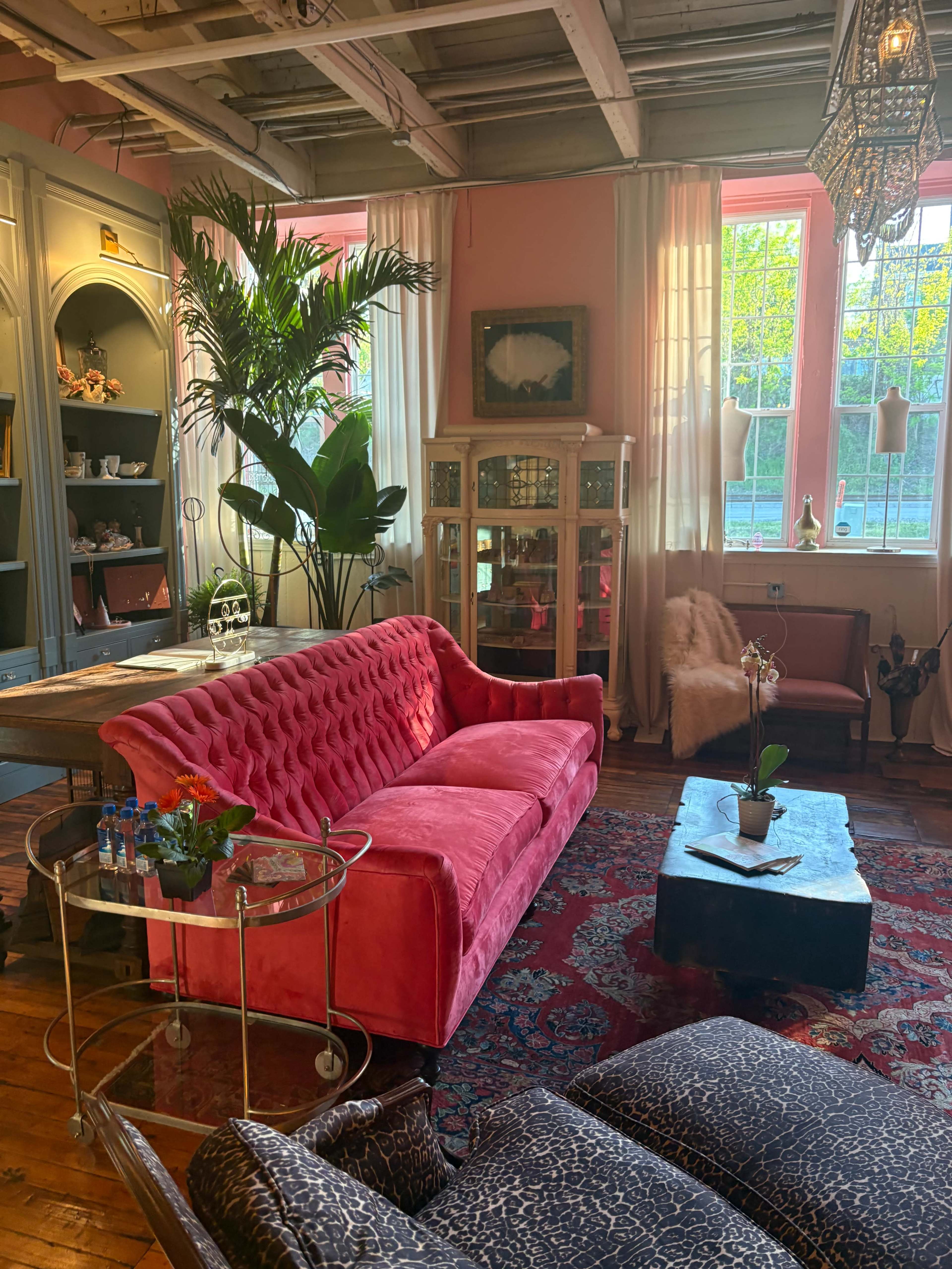The image shows a cozy room featuring a pink tufted sofa, a glass side table with refreshments, a decorative cabinet, and large windows allowing natural light to enter.
