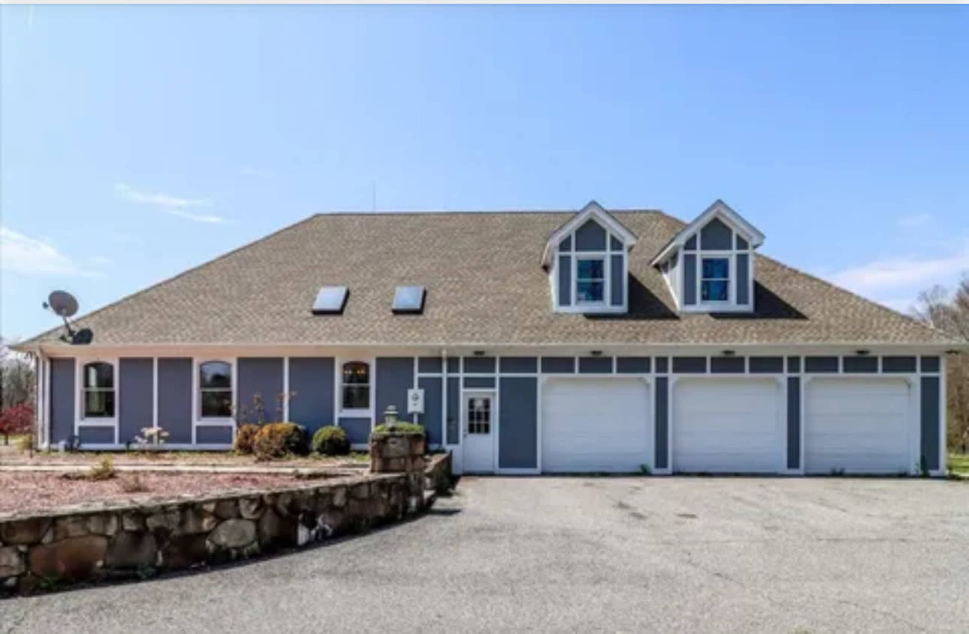 A large, two-story house with a gray exterior and a stone-bordered driveway features a central entrance flanked by garage doors.