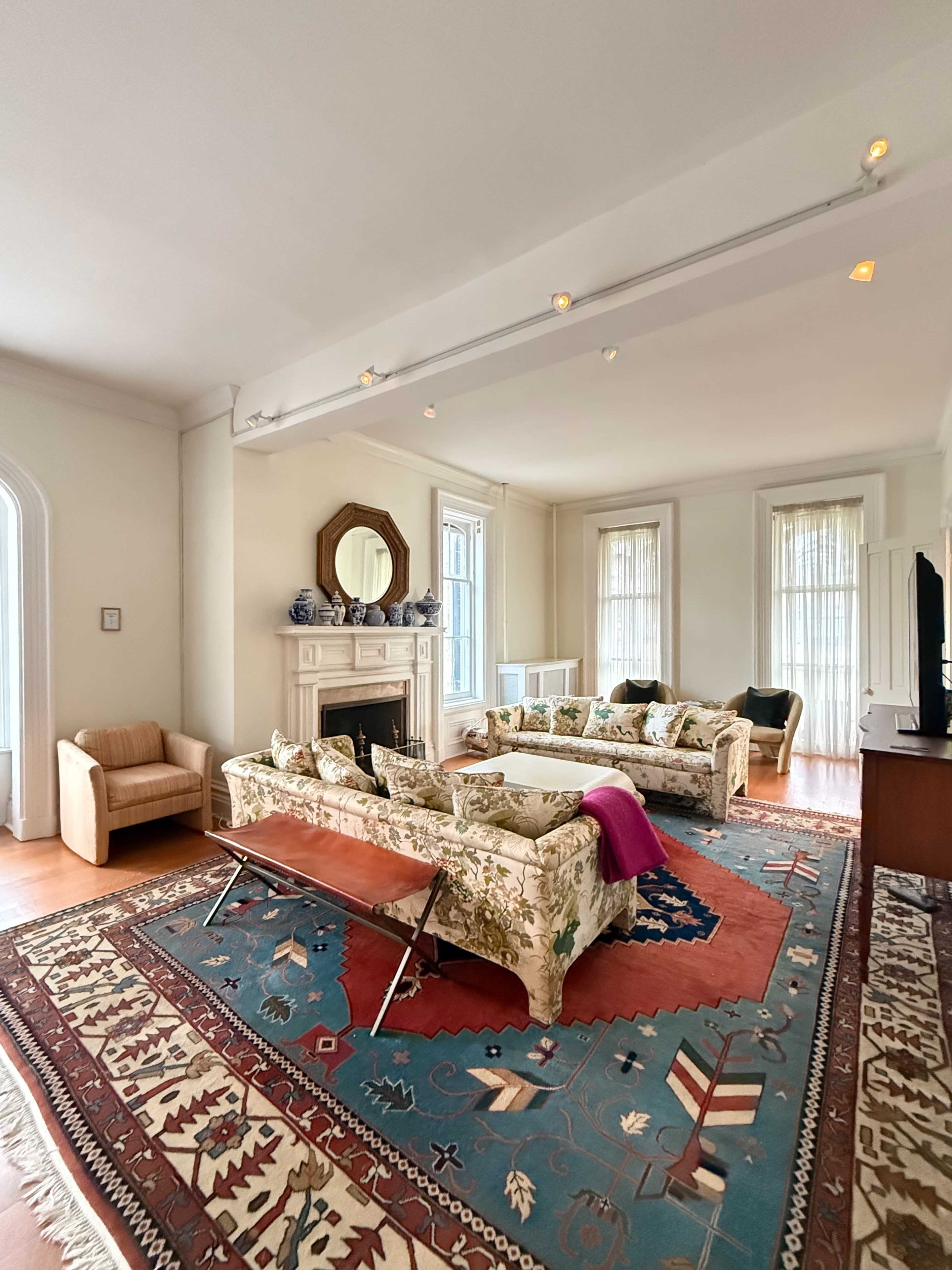 The living room features two floral-patterned sofas, a wooden fireplace with a mirror above it, and a large area rug with geometric designs.