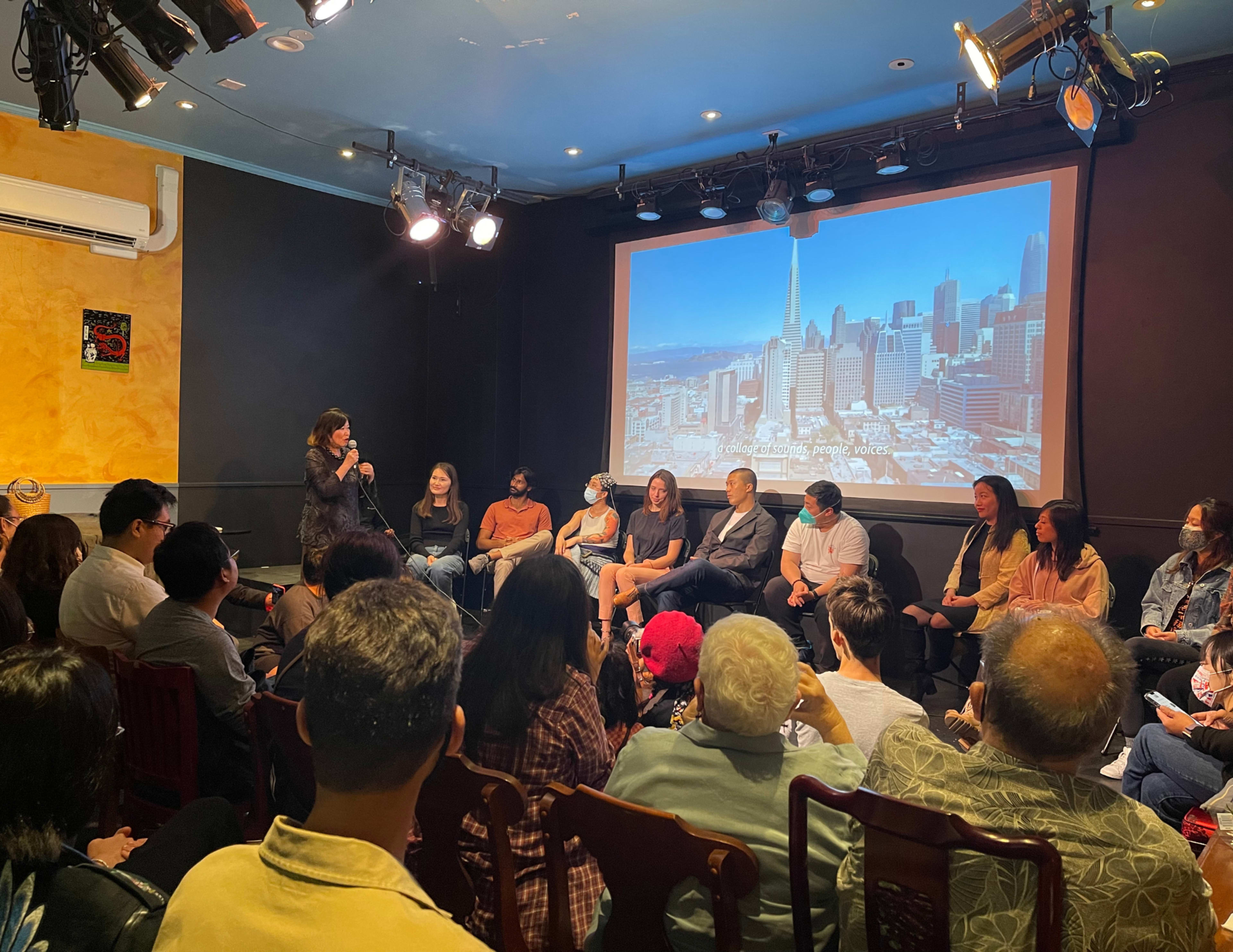 A panel discussion takes place on stage in front of an audience, with a city skyline displayed on a screen behind the speakers.